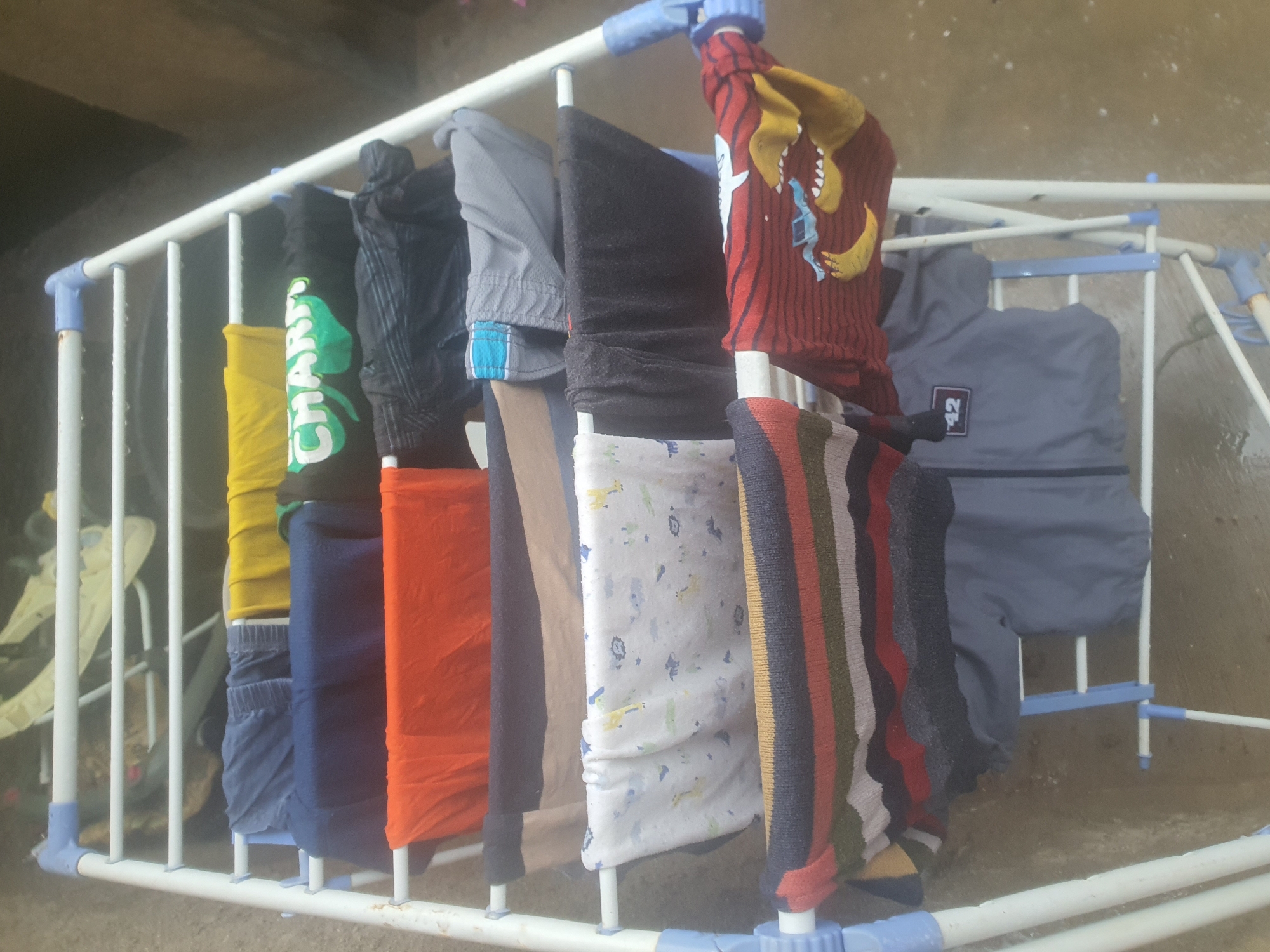 The image shows a white metal clothes drying rack with various items of clothing spread across its horizontal bars. The clothing includes blue trousers, a yellow t-shirt, a green t-shirt with white lettering that appears to say "CHARM", an orange t-shirt, dark-colored trousers or leggings, gray shorts with a blue waistband, a white fabric with a pattern of small animals, dark fabric that appears to be pants, a striped sweater with maroon, gray, and white stripes, and a gray jacket with a black patch displaying the number "42". The drying rack is set up outdoors on a wet, uneven concrete surface. The background is blurred but suggests an outdoor, possibly urban or semi-urban, environment. The overall lighting indicates it is daytime. This scene is consistent with laundry being dried outdoors in Jos, Nigeria, where such drying racks are common in residential areas.