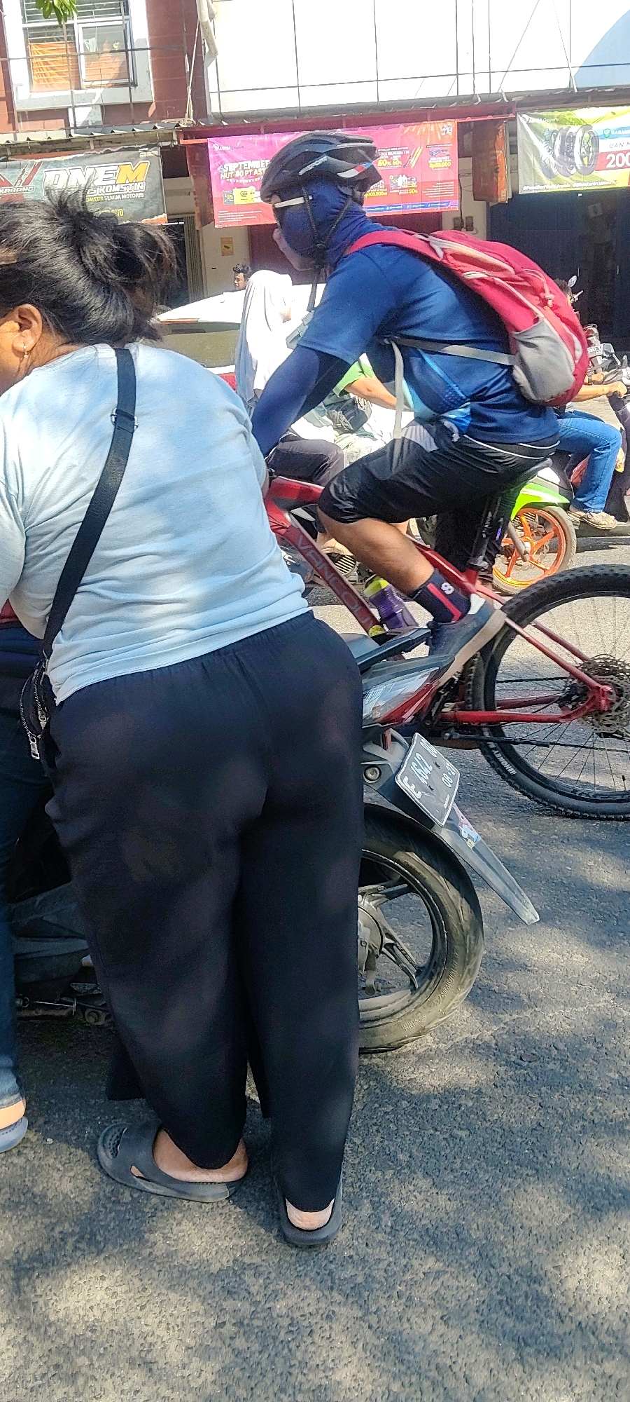 The image shows a street scene in Cibatu, Indonesia, with multiple people and vehicles. In the foreground, a person is wearing a light blue t-shirt and black pants, with a black bag slung over their shoulder. This individual is facing away from the camera and appears to be standing next to a motorcycle. To the right, another person is riding a bicycle, wearing a blue long-sleeved shirt, black shorts, a helmet, and a red backpack. This cyclist is also wearing a blue face covering. The background reveals a street with buildings and advertisements. A green motorcycle is parked to the right of the cyclist, and another motorcycle is visible further in the background. The scene is illuminated by daylight, casting shadows on the asphalt road.
