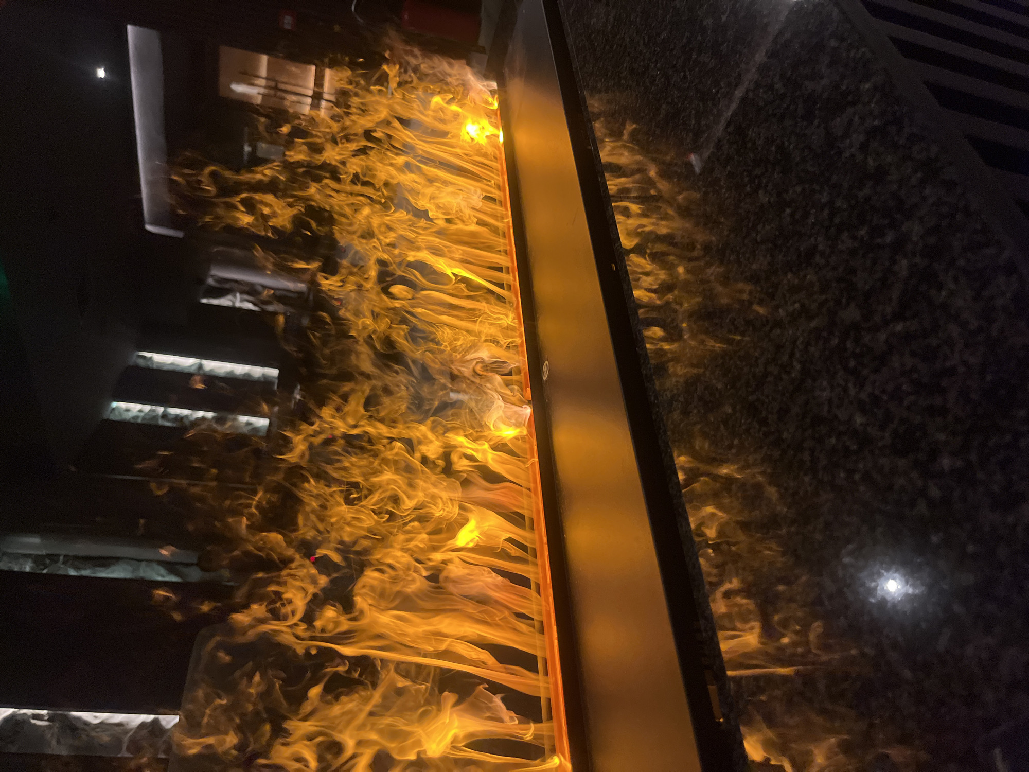The image shows a decorative artificial fire installation within an interior space. The "flames" are illuminated in shades of orange and yellow. The installation appears to be encased in a vertical structure with dark, possibly granite, side panels. A set of stairs is visible behind the installation, reflecting the artificial light. Light fixtures are visible in the reflected surfaces. The location is likely Sofia, Bulgaria.