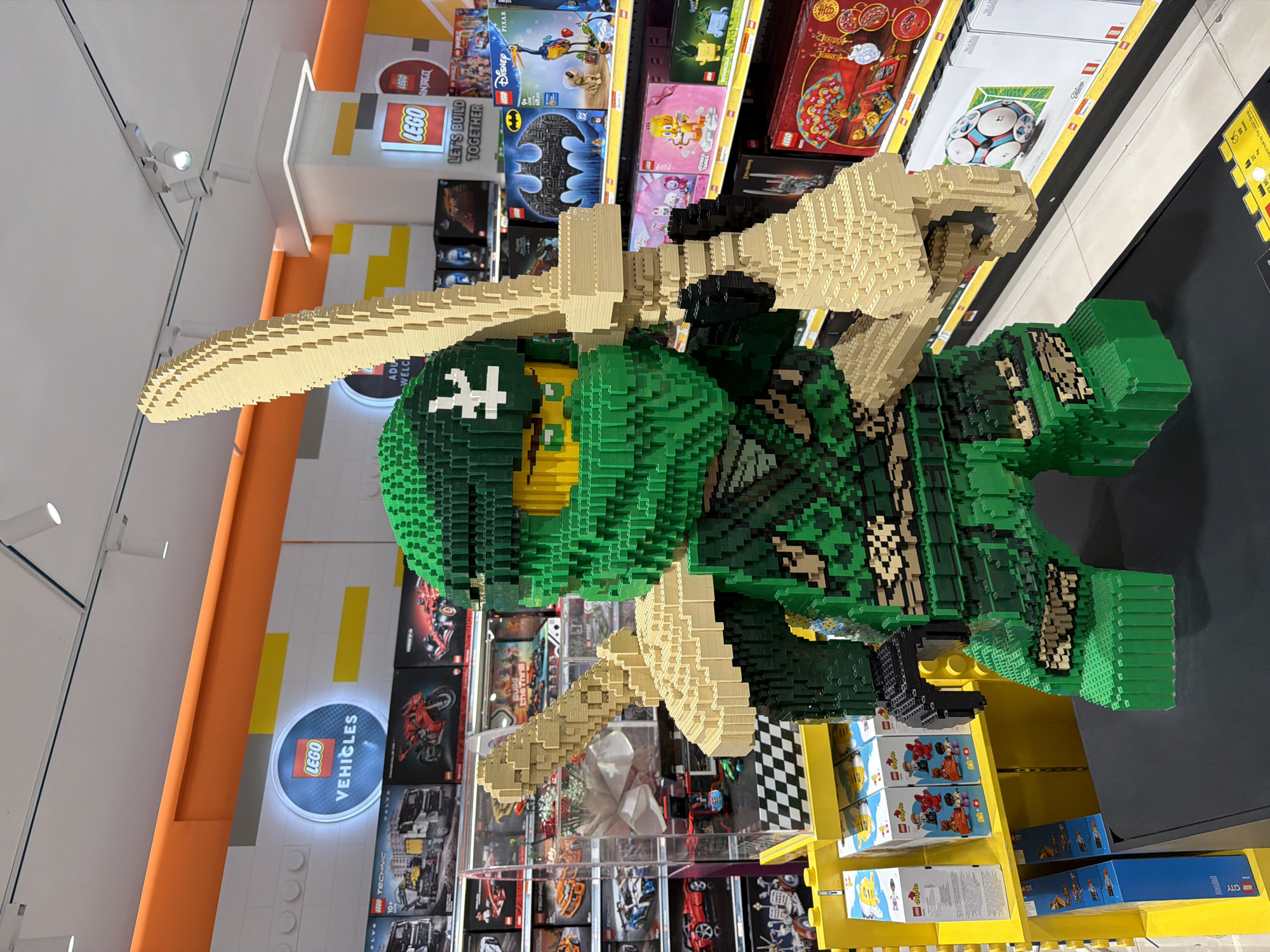 The image captures a life-sized Lego sculpture of a Ninja Turtle character in a retail environment. The sculpture is primarily composed of green and tan Lego bricks. The turtle's head and mask are green, with a white cross symbol on the mask. The two tan katanas are mounted to the Lego turtle's back.
Behind the Lego sculpture, there are shelves filled with Lego sets, including Lego Vehicles, Lego Ninjago, Lego Disney, and other Lego themes. The shelves are organized with the Lego boxes facing forward to display the product packaging.
The color scheme of the surrounding walls and shelving is a mix of white, yellow, and orange, characteristic of Lego retail environments. The environment appears to be well-lit.
No people are directly visible in the frame. There is no explicit indication of being in Kuta, Indonesia.