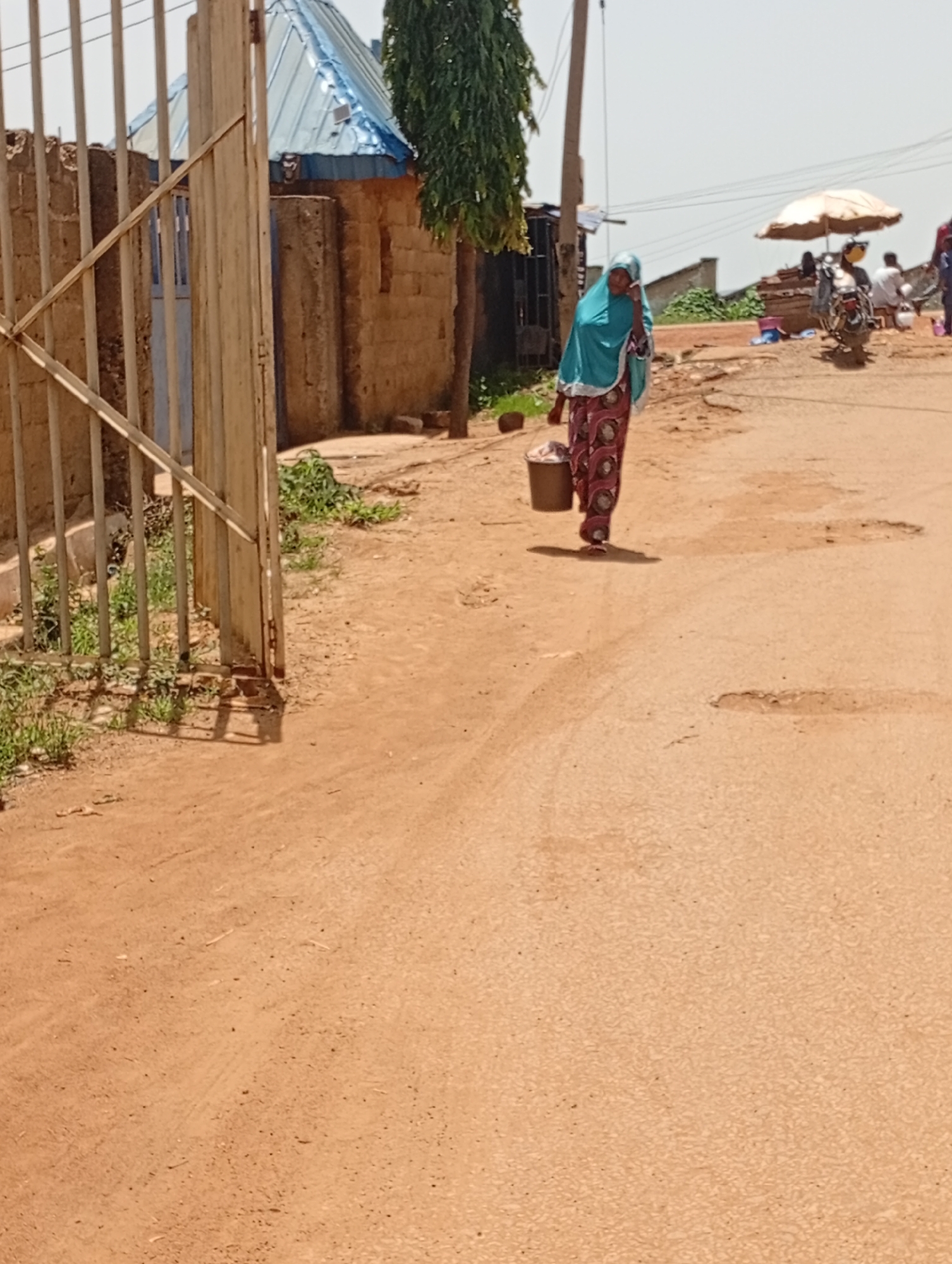 In Rafin Par, Nigeria, a woman wearing a turquoise headscarf and a patterned dress walks down a dirt road, carrying a brown bucket in her right hand. The road is uneven with potholes and debris. To the left, a metal gate with vertical bars stands open, revealing a courtyard with a brick building and a blue corrugated metal roof. Vegetation, including grass and a tall, thin tree, grows along the roadside and near the gate. In the background, a motorcycle with an umbrella canopy is parked, and other people are visible in the distance. The sky is bright and clear.