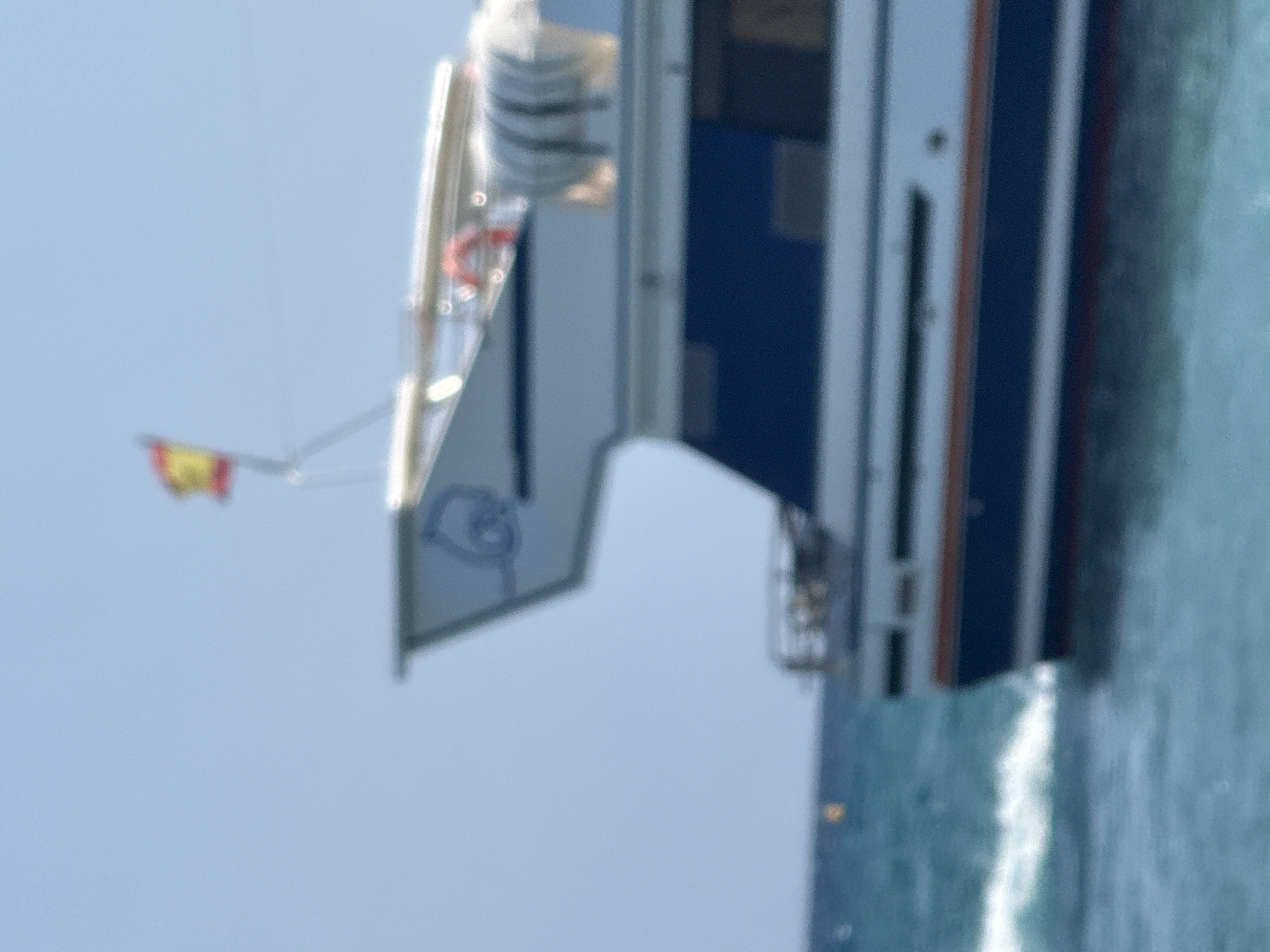 The image shows the stern of a white and blue boat sailing on the Mediterranean Sea. A Spanish flag is visible on a mast at the top of the boat. The water is choppy and appears to be a bright turquoise color. The sky is clear and blue. The boat is white with a dark blue stripe running along its hull and a red stripe above the blue stripe. There is a logo on the side of the boat that appears to be a blue outline of a dolphin. In the distance, a small yellow buoy is visible in the water. The overall scene suggests a sunny day at sea, likely off the coast of Blanes, Spain, given the Spanish flag.