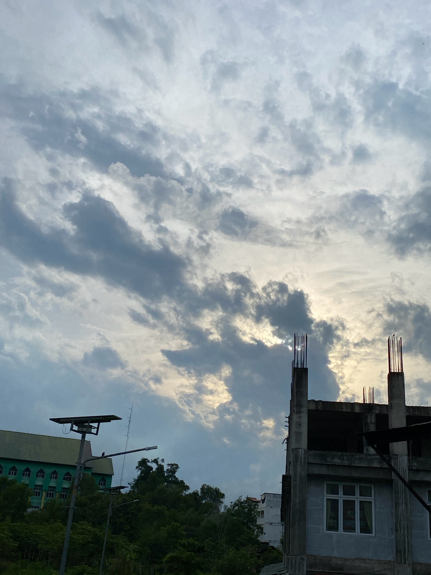 The image depicts a cloudy sky over a developing urban area. In the foreground, there is a partially constructed concrete building with exposed rebar protruding from the tops of concrete pillars, suggesting ongoing construction. Further to the left, a green building with a distinctive arched window design is visible, likely a public facility or institution. A solar-powered street lamp stands prominently in front of this green building. Dense green foliage covers the lower slopes of the terrain, with trees and other vegetation obscuring the base of the structures. The overall scene suggests a moment in time during the day, with the sun partially visible through the overcast sky. The presence of construction, residential or institutional buildings, and natural elements indicate a typical urban landscape in Lhokseumawe, Indonesia.