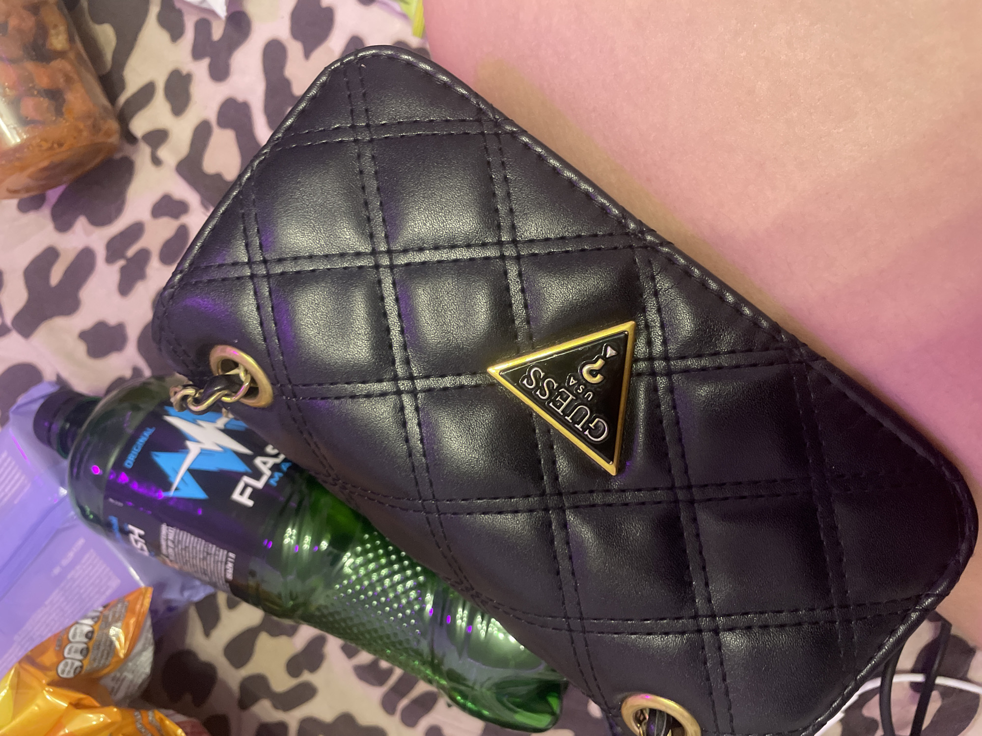 A black quilted clutch purse with a gold Guess logo is resting on a person's leg. To the left of the purse is a green bottle with the word "FLASH" written on it. The background consists of a leopard-print fabric and some other objects that are out of focus, possibly snacks. The scene appears to be indoors, and the lighting suggests it might be evening or a dimly lit room. There is no indication of the city of Shcheglovo, Russia.