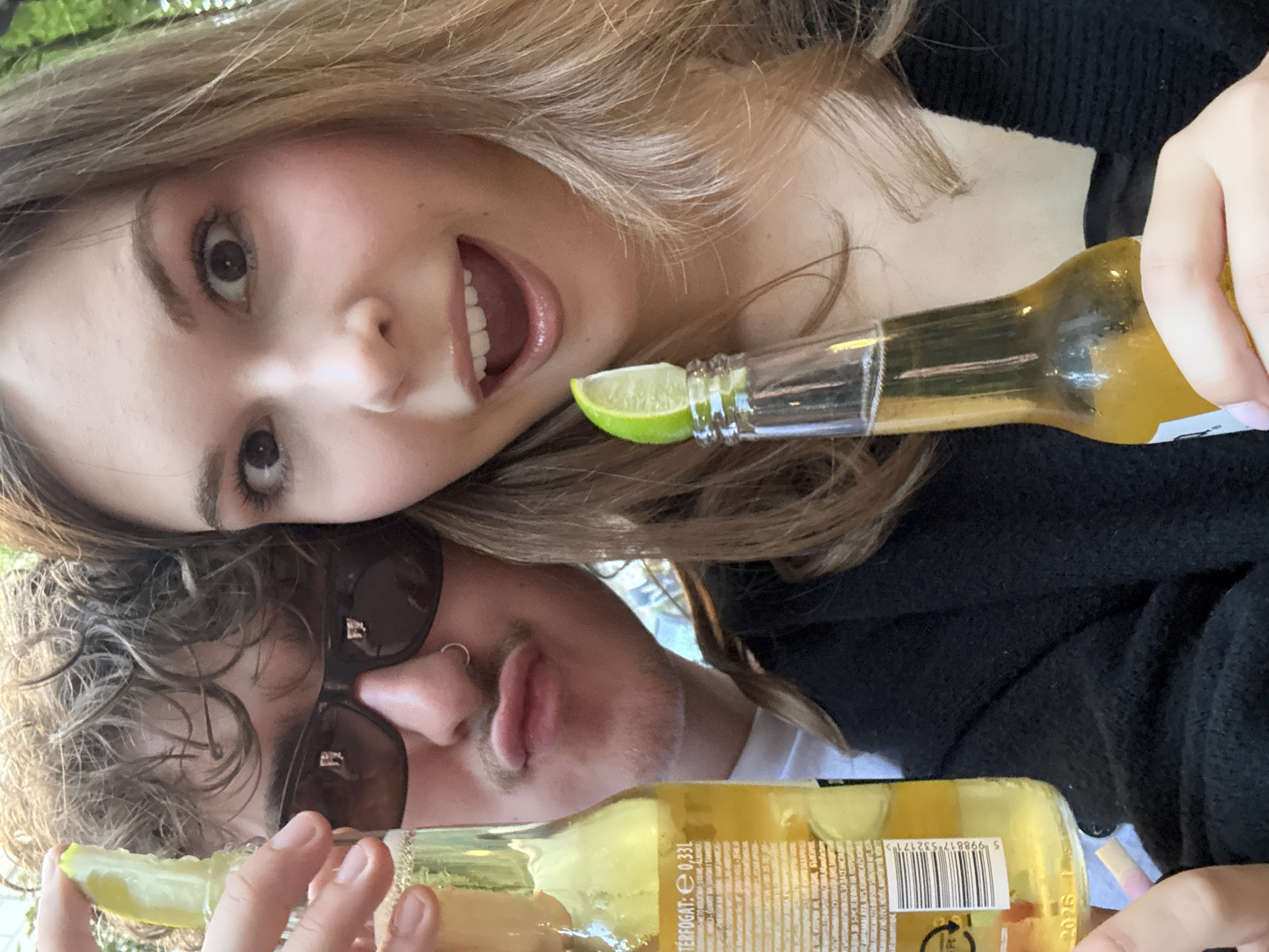 Two individuals, a male and a female, are taking a selfie outdoors. The male, wearing sunglasses and a nose ring, is holding a bottle of beer with a lime wedge in it. He has curly brown hair and is making a puckered expression. The female, with long blonde hair, is also holding a bottle of beer with a lime wedge. Her eyes are wide and her mouth is open, as if she is surprised or excited. The background suggests a casual outdoor setting, possibly a park or a beer garden. The lighting indicates it is daytime. The people appear to be enjoying themselves, possibly at a social gathering or a casual outing. The specific location in Belgrade, Serbia, cannot be determined from the visual information provided.