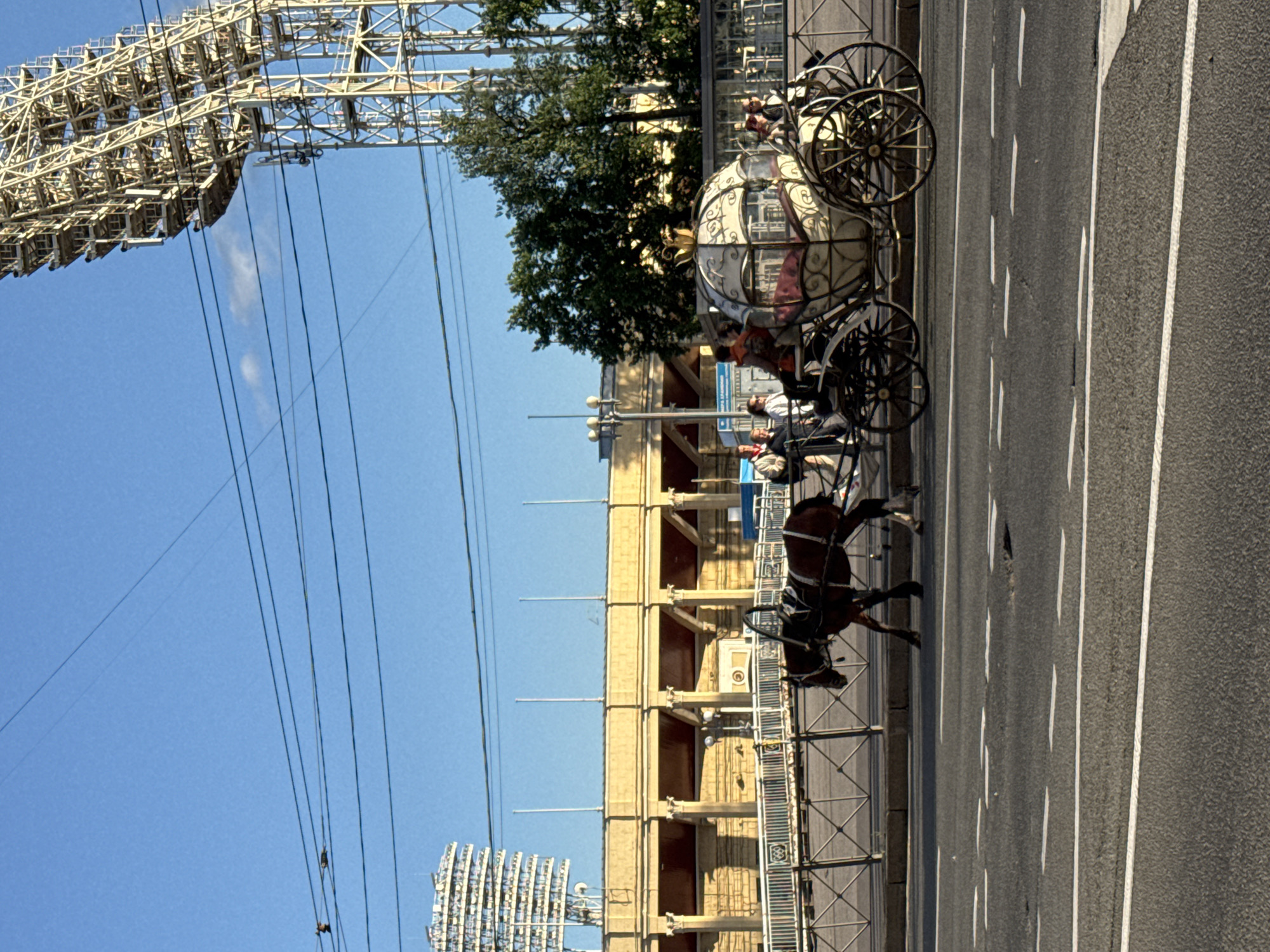A horse-drawn carriage, designed to resemble a Cinderella-style coach, travels along Nevsky Prospekt in Saint Petersburg, Russia, on a clear, sunny day. The carriage is ornate, with white and gold detailing and a decorative crown on top. It carries several passengers, including women and children. A brown horse pulls the carriage. In the background, a light yellow, neo-classical building with columns and arches lines the street, and further behind, a large stadium-like structure with floodlights is visible against the bright blue sky. Overhead power lines crisscross the scene. The street in the foreground is paved asphalt with white lane markings, showing some signs of wear and tear. A metal fence separates the sidewalk from the roadway.