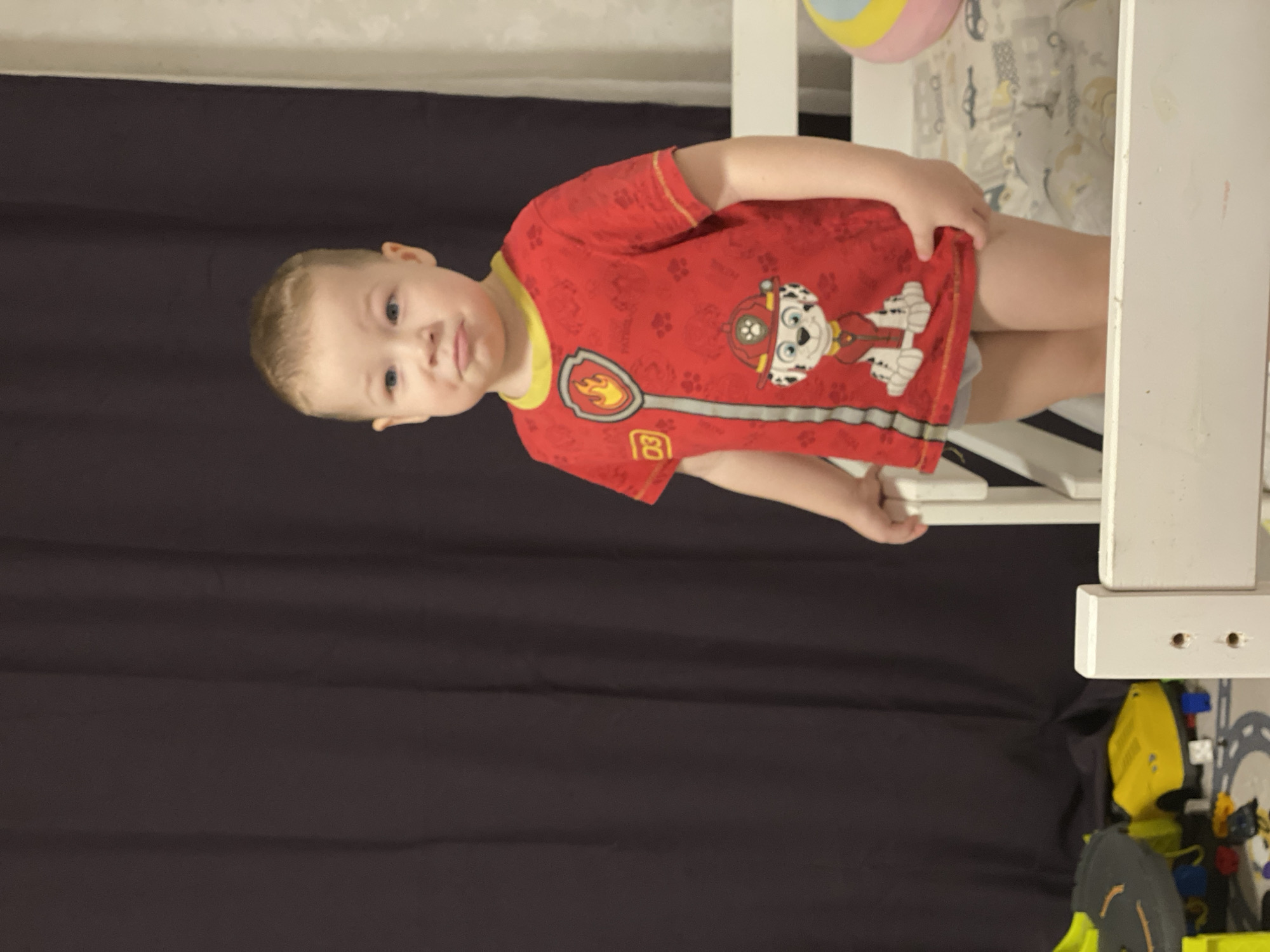 A young child is depicted in a close-up, indoor setting. The child, appearing male with short, light-colored hair and blue eyes, is standing and looking directly at the viewer with a slight pout. He wears a red short-sleeved t-shirt featuring a yellow collar and sleeve trim. The t-shirt has a graphic print depicting a flame badge, the number "03," and the character Marshall from Paw Patrol, a white dalmatian in a firefighter uniform. Light-colored underwear or a pull-up is visible beneath the t-shirt. The child's left hand rests on a white wooden bed frame or crib railing that partially obscures the lower portion of his body. His right hand is near the hem of his t-shirt. Behind the child and to the right, a bed is visible with a light-colored sheet patterned with vehicle outlines, and a multi-colored spherical toy (yellow, blue, pink) rests on it. To the lower left, various toys, including a yellow toy car, a dark grey toy, and colorful building blocks, are scattered on the floor. The background consists predominantly of a dark brown curtain on the left and center, with a section of a light-colored wall visible on the far right. The specific location within the city of Sochi, Russia, cannot be determined from the visual information provided in the image.