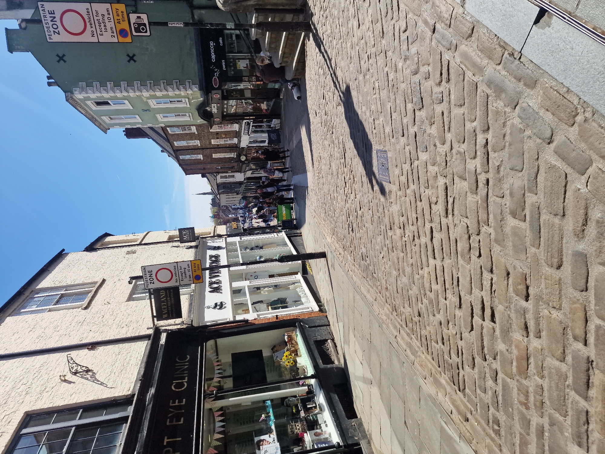The image depicts a street scene in Durham, United Kingdom, characterized by cobblestone paving and a mix of historic buildings housing various businesses. The street appears to be a pedestrian zone, evidenced by signage.
On the left side of the frame, a building with a shop front labeled "OPT EYE CLINIC" is visible. Next to it, is "WACELAND" and "ACE VINTAGE" A sun dial is fixed to the wall above.
Further down the street, the view opens up, revealing more shops and what appears to be a restaurant.
On the right side of the frame, the cobblestone street is featured prominently, with its uneven surface and visible seams. Shadows are cast upon it.
People are walking along the street, and a general sense of daytime activity is conveyed.
Signs indicate parking restrictions and pedestrian zones.