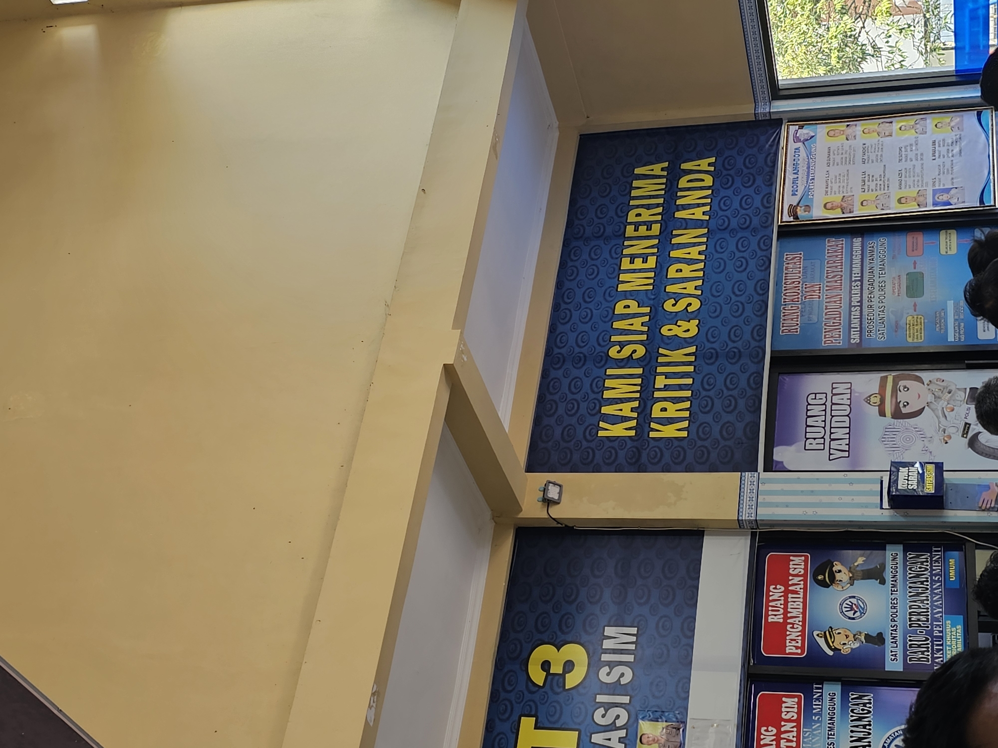 The image shows a section of a building with several signs and posters. In the foreground, there are people, suggesting an indoor public space. The prominent signage includes "T 3 ASI SIM" on a blue background with circular patterns, and to its right, a large blue banner with yellow text that reads "KAMI SIAP MENERIMA KRITIK & SARAN ANDA" (We are ready to accept your criticism & suggestions). Below these main signs, further posters are visible, including one for "RUANG PENGAMBILAN SIM" (SIM Collection Room) with cartoon figures of police officers and another for "RUANG YANDUAN" (Service Room) with a similar cartoon. To the far right, a "PROFIL ANGGOTA POLRES TEMANGGUNG" (Member Profile of Temanggung Police) poster is displayed, featuring photos and names of police personnel. The overall environment appears to be a public service area, likely a police station or a government office, given the nature of the signage. The location is identified as Madureso, Indonesia.