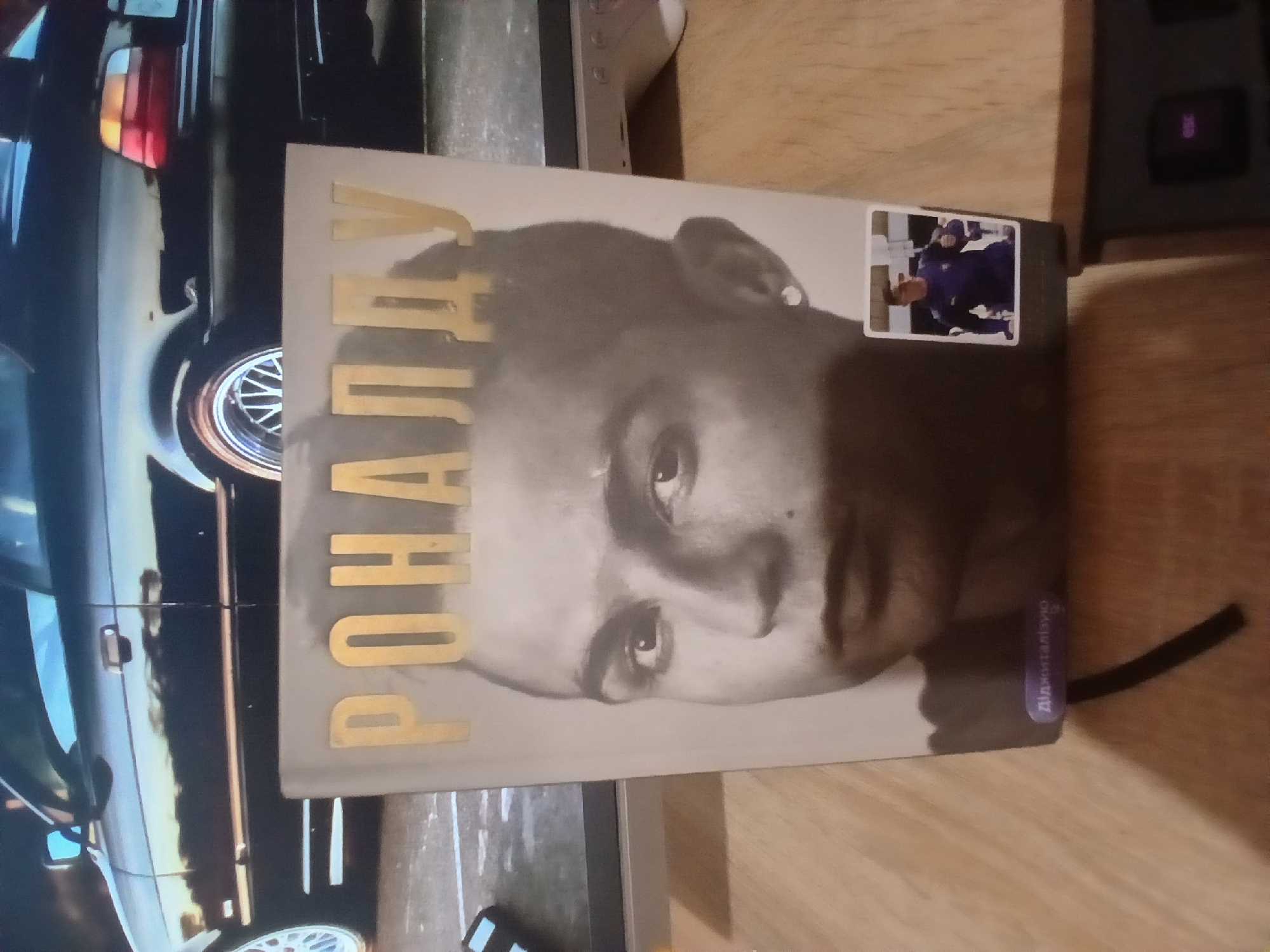 The image shows a book with "РОНАЛДУ" (Ronaldo) written in gold letters on the cover, featuring a portrait of Cristiano Ronaldo. The book is placed on a wooden surface, with a portion of a dark-colored car visible in the background, specifically its rear end with taillights and a wheel. A small inset image of Cristiano Ronaldo in a dark blue athletic outfit is affixed to the cover. The environment appears to be indoors, with a desk or table in the foreground and a monitor or TV screen behind the car. There is no identifiable information suggesting the location is in the city of Dnipro, Ukraine.