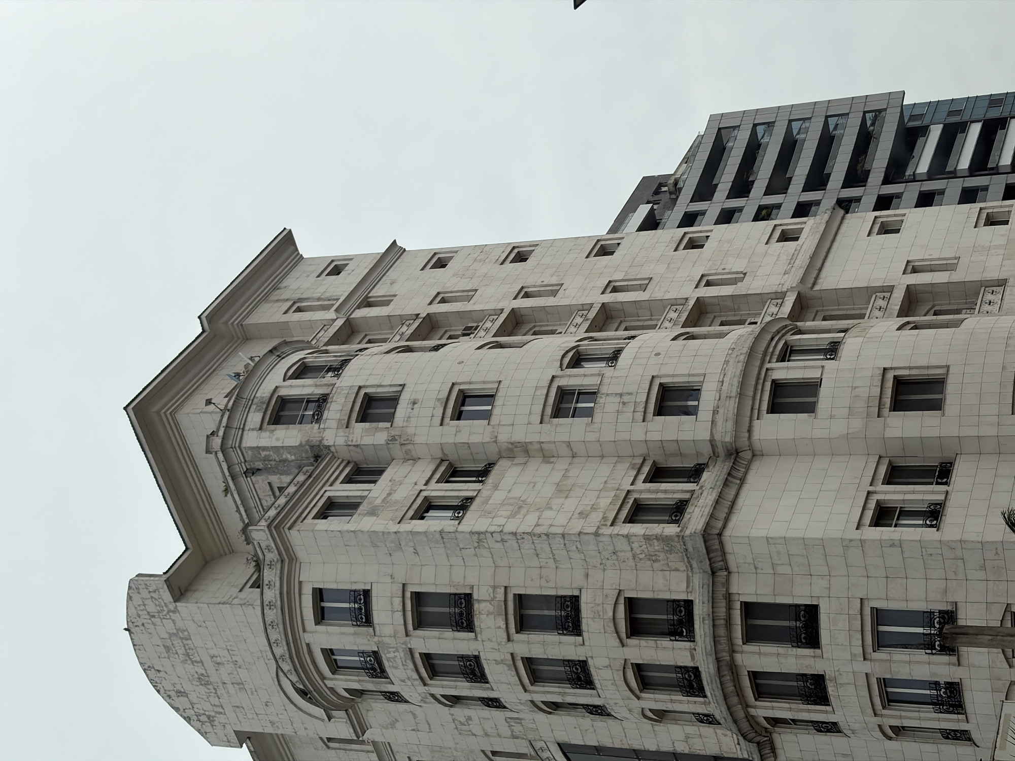 An architectural shot, taken from a low angle, features a distinctive multi-story building under an overcast sky in Falomo, Nigeria. The primary subject is a light-colored building, likely constructed from stone or concrete, exhibiting a classical or Art Deco-inspired design. Its facade is characterized by a grid-like pattern of square tiles, numerous rectangular windows, and a prominent curved section that adds a dynamic element to its structure. Many windows are adorned with intricate black wrought-iron railings, adding a decorative touch. The building features ornate cornices, moldings, and ledges, particularly at its upper levels and corners, suggesting a rich architectural heritage. Some areas of the facade show signs of weathering or age.
Behind and to the right of this main structure, a more modern, dark-colored building is partially visible. This secondary building contrasts sharply with the foreground structure, featuring a contemporary design with glass panels and strong geometric lines, possibly indicative of a newer construction or renovation.
The scene depicts an urban setting, with no human activity or specific event taking place. The sky is uniformly gray and overcast, suggesting a cloudy day with diffused light. There is no visible text in the image. The overall impression is one of architectural contrast within a bustling city environment.