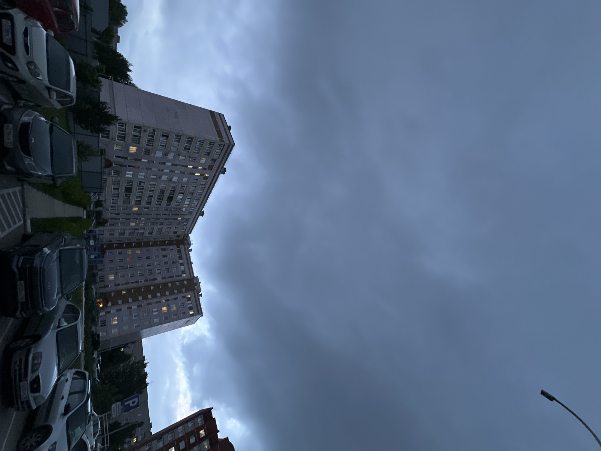 The image displays a residential street scene in Chaikovsky, Perm Krai, Russia, observed during evening twilight or under heavily overcast conditions. The upper two-thirds of the frame are filled with a dark, uniform grey, heavily clouded sky, with a partial view of a dark street light fixture curving into the upper right corner.
Dominating the lower and middle ground is a large, multi-story apartment building, distinctive for its angled facade on the left side, giving it a trapezoidal profile at its base. The building features numerous rectangular windows, many of which are illuminated with warm yellow light, indicating occupancy. Its exterior is light grey or off-white, with horizontal bands of a light brown or beige color near the roofline and between some floors. To the far right, a partial view of another multi-story building, appearing darker in tone (possibly red brick), is visible, also with illuminated windows.
In the foreground and middle ground, a paved parking area is visible, occupied by several parked passenger vehicles. From left to right, partially visible, is a red car. Next to it is a light-colored hatchback with Russian license plate "A366KA 59". To its right, a dark grey or black sedan bears license plate "H990CA 59". Further back, a silver SUV with license plate "B433BM 59" is parked, along with another silver sedan. A blue rectangular parking sign with a white "P" is visible in the middle-right background.
The ground features a narrow paved walkway or sidewalk running alongside the main building, bordered by low green shrubs and dark fencing. Mature trees and additional greenery are visible behind the parked cars and in the background, obscured by the dim light. No individuals are visible in the scene. The overall lighting is dim, reflecting the overcast sky and the time of day.