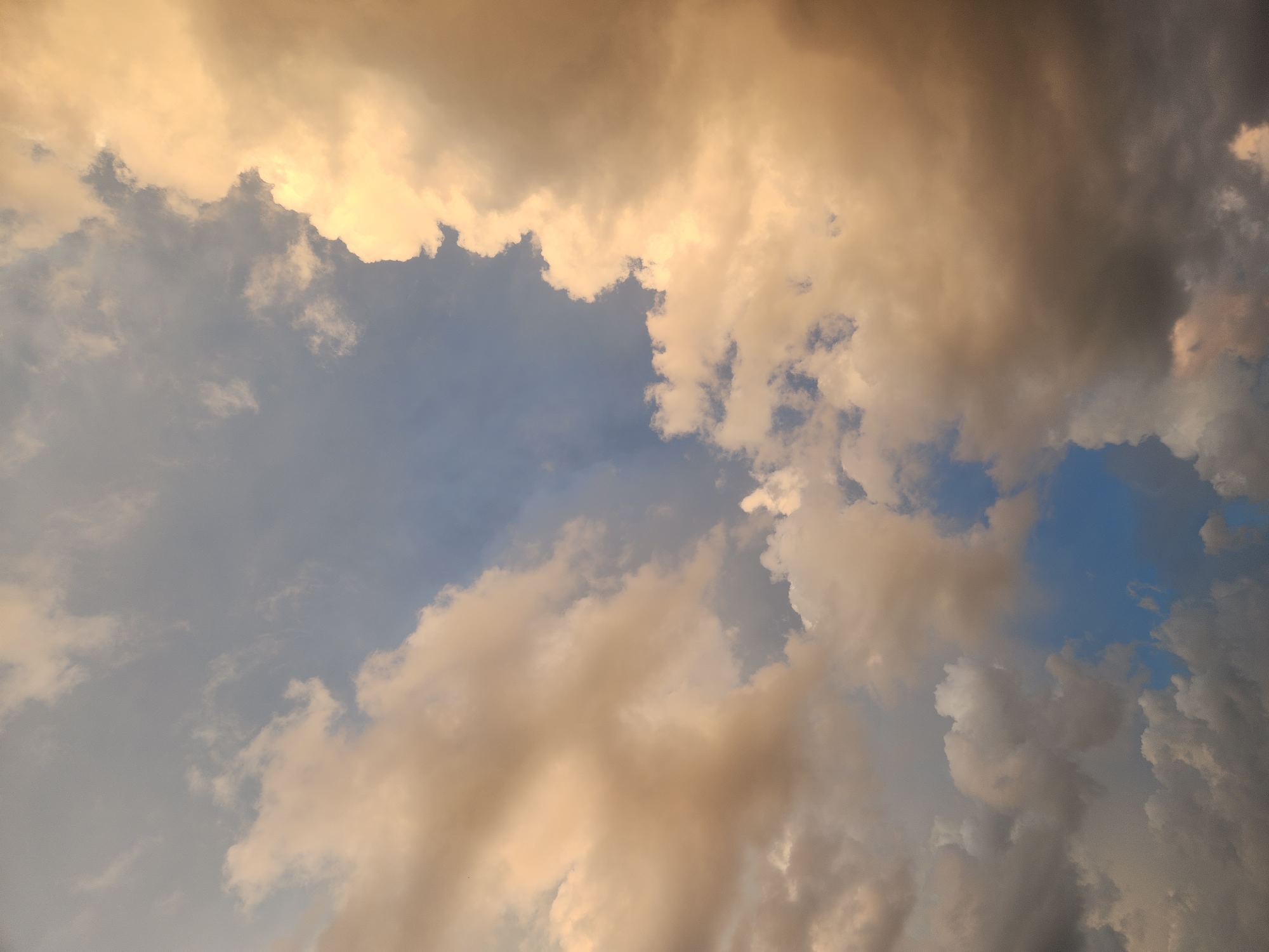 The image depicts a cloudy sky with patches of blue visible. The clouds are illuminated by warm, golden light, suggesting either sunrise or sunset. The clouds vary in color from light tan and cream to darker grays and blues, indicating different densities and depths. The overall scene is abstract, focusing on the texture and light play of the sky. There are no discernible landmarks or objects to indicate a specific location within Winter Park, United States of America, nor are there any people or actions depicted.