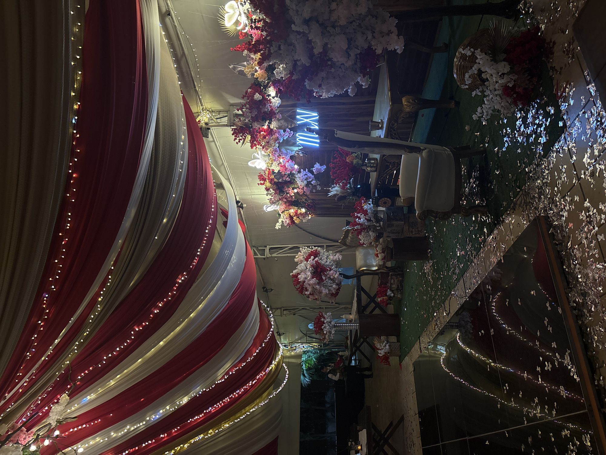 The image shows an interior event space in Chilpancingo, Mexico, decorated for a celebration, possibly a wedding. Red and white draped fabrics with string lights adorn the ceiling and walls. Floral arrangements with red and white flowers and greenery are also present. Furniture, including white chairs, are arranged around tables. Silver confetti scatters the floor, part of which is mirrored. A neon sign reading "m v" hangs amongst the flowers.
