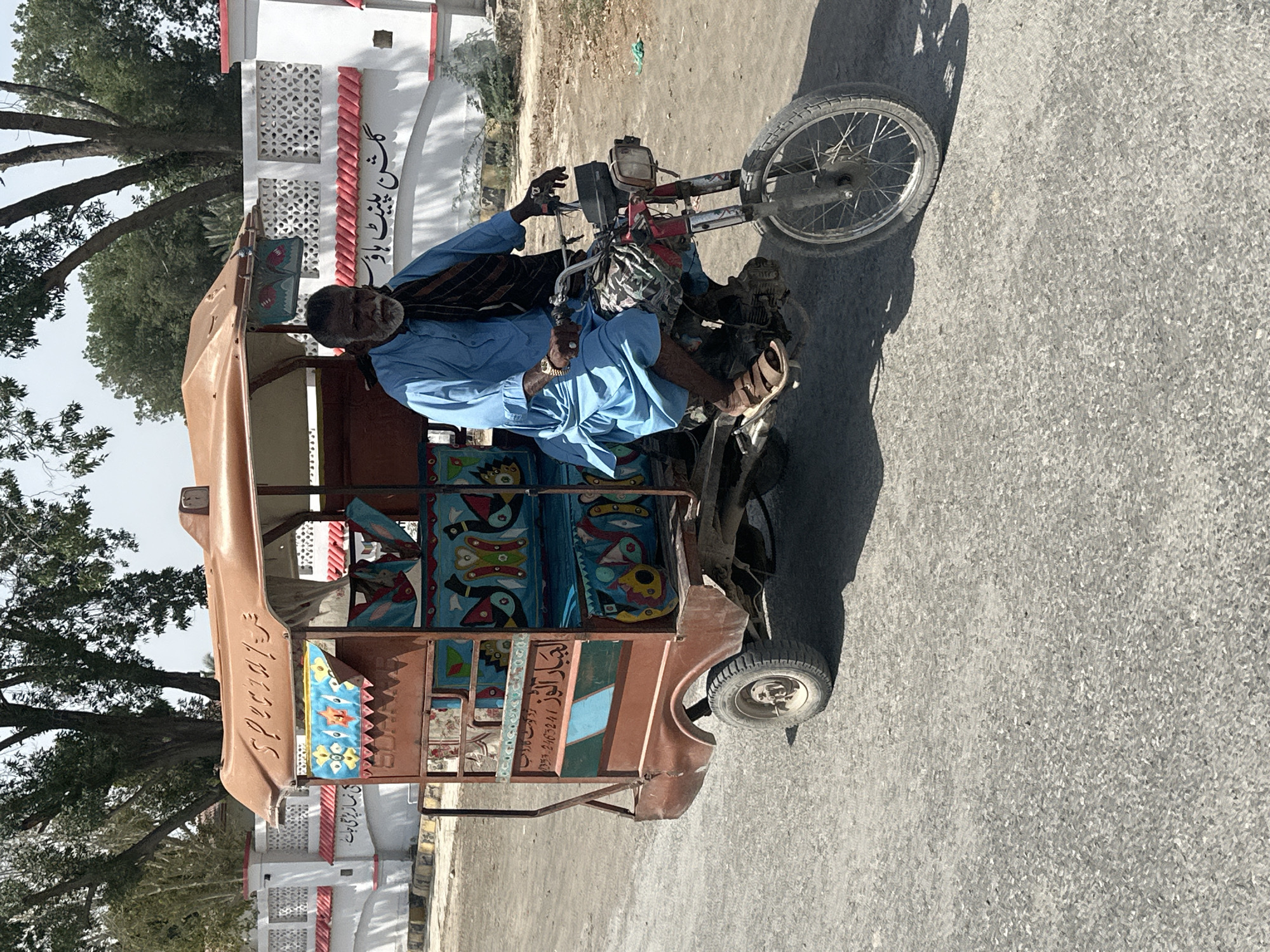 The image, rotated 90 degrees counter-clockwise, captures an older man driving a colorfully decorated motorized rickshaw on a paved road. The man has a dark complexion, a salt-and-pepper beard, and is wearing a light blue shalwar kameez-style outfit with a dark, possibly striped, shawl draped over his shoulders. He wears a gold-colored watch on his left wrist and light-colored sandals. He is seated in the driver's position, holding the handlebars.
The three-wheeled rickshaw is predominantly brown but features elaborate, multi-colored folk art designs on its passenger compartment, including stylized birds, fish, and geometric patterns in shades of blue, red, yellow, and green. The vehicle appears to be a common type of auto-rickshaw or 'chingchi'.
The scene is brightly lit by the sun, casting strong shadows on the road, indicating a clear day, likely morning or late afternoon. In the background, there are large green trees to the left and a white building with red decorative elements and patterned cutouts to the right. The road surface appears to be asphalt, with some dry, dusty areas nearby.
Visible text includes "گمن پشٹ ہاوس" on the building. On the rickshaw, the English word "SPECIAL" is seen, along with Urdu script: "اسپیشل" (Special), "عبدالرحمن" (Abdul Rahman), a phone number "0333 2463241", and "اللہ نظر بد سے بچائے" (May Allah protect from the evil eye).