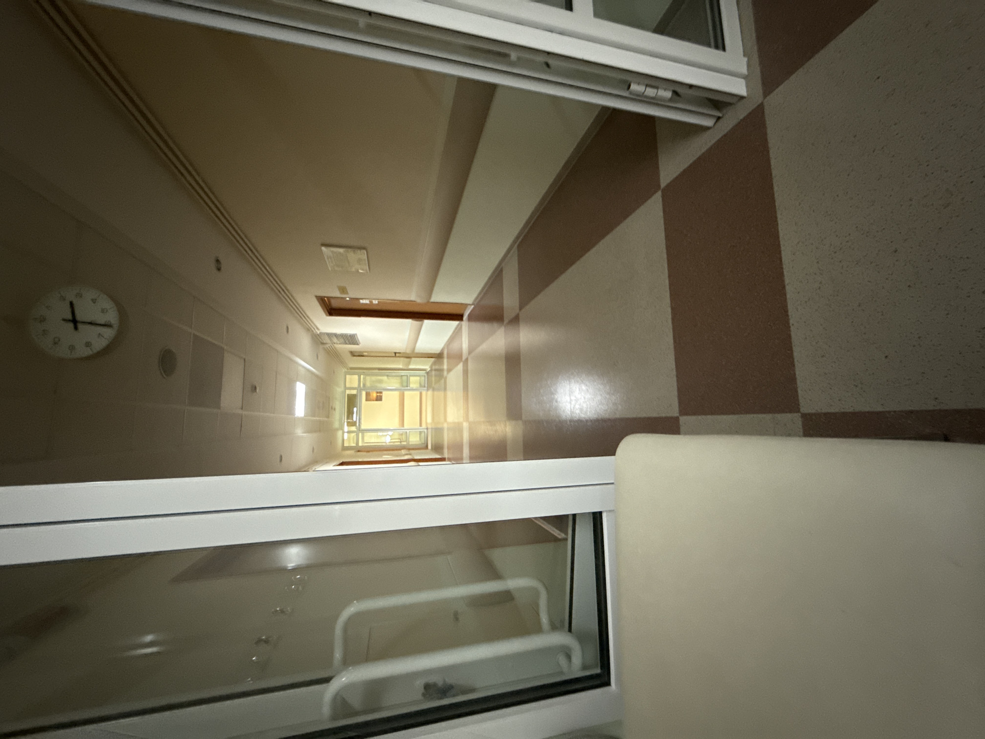 The image captures a long, narrow, empty hallway or corridor, viewed from an elevated perspective looking downwards, possibly from an upper floor or balcony through a window or railing. The foreground is dominated by a white window frame and glass, reflecting white railings and other indistinct elements from the immediate vicinity of the camera. To the right in the foreground, a cream-colored, soft-looking object, possibly part of a chair or bed, is partially visible.
The hallway features light beige walls with horizontal and vertical decorative bands, some darker brown, creating a checkered or striped pattern, particularly on the right wall. The floor also exhibits a checkered pattern of light and dark brown tiles. The ceiling is plain and light-colored, with rectangular light fixtures and vents sparsely placed along its length. A round wall clock is visible on the left wall, showing the time around 10:45.
Further down the hallway, a dark wooden door frame is visible, and the perspective converges towards a brightly lit area at the far end, suggesting windows or a more intensely lit section. The overall lighting is artificial and uniform, creating a somewhat sterile, institutional atmosphere. There are no people present, and no activity is taking place. The scene suggests a public or semi-public building, such as a hospital, school, or office complex, in Moscow, Russia.