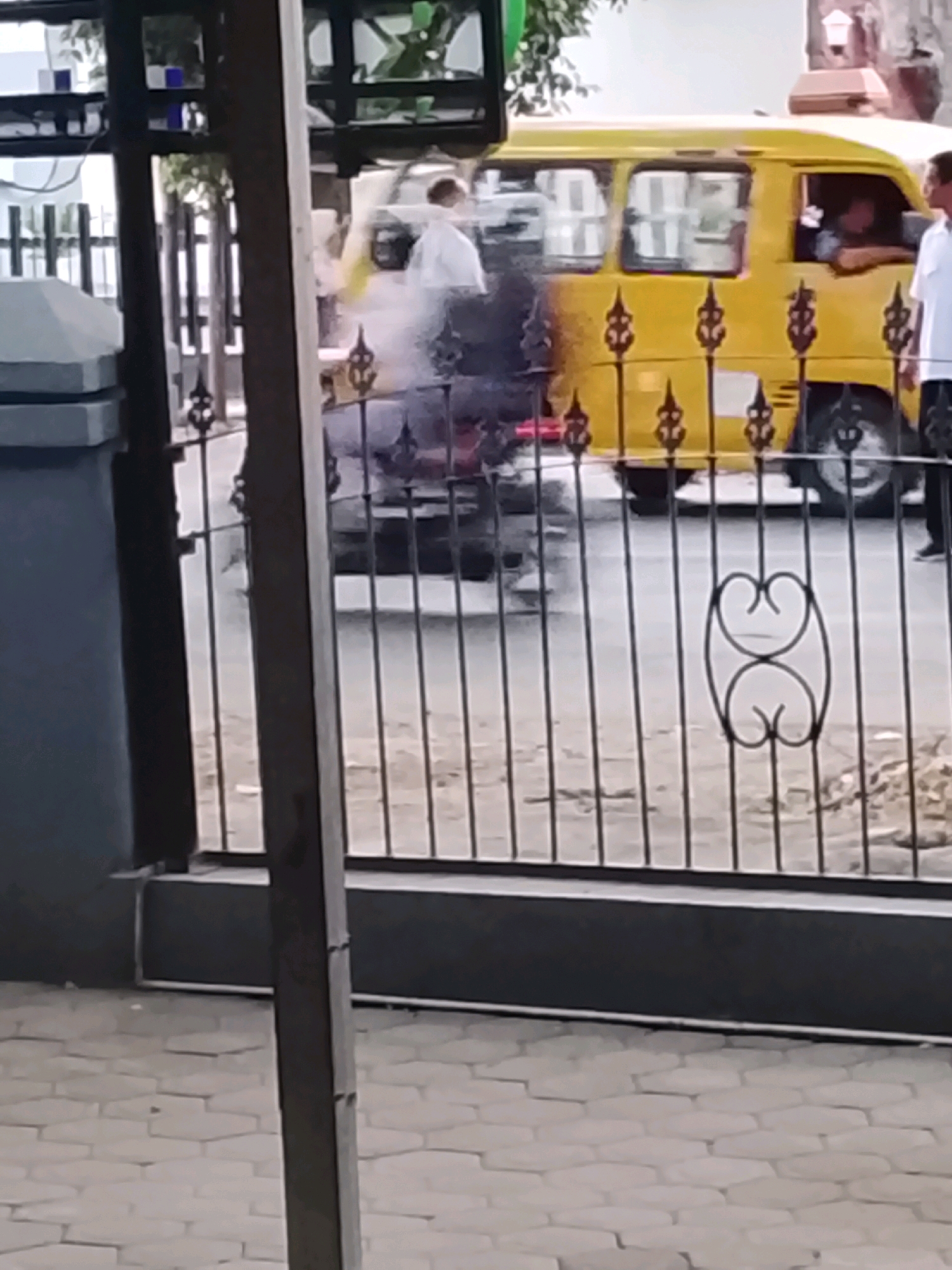 The image captures a street scene in Slawi, Indonesia, dominated by a yellow van. The van, with its windows reflecting the surroundings, is parked on a paved road. A person, appearing to be a male wearing a white shirt, is leaning out of the driver's side window, and another person in a white shirt is visible near the front passenger side of the van, partly obscured by smoke emanating from the vehicle. The smoke suggests a potential issue, such as a mechanical problem or an accident. A decorative black metal fence runs across the foreground, partially obstructing the view of the van and the figures. Beyond the fence, other vehicles and elements of a typical urban environment are discernible, though less sharply focused. The ground in front of the fence is paved with hexagonal stones.