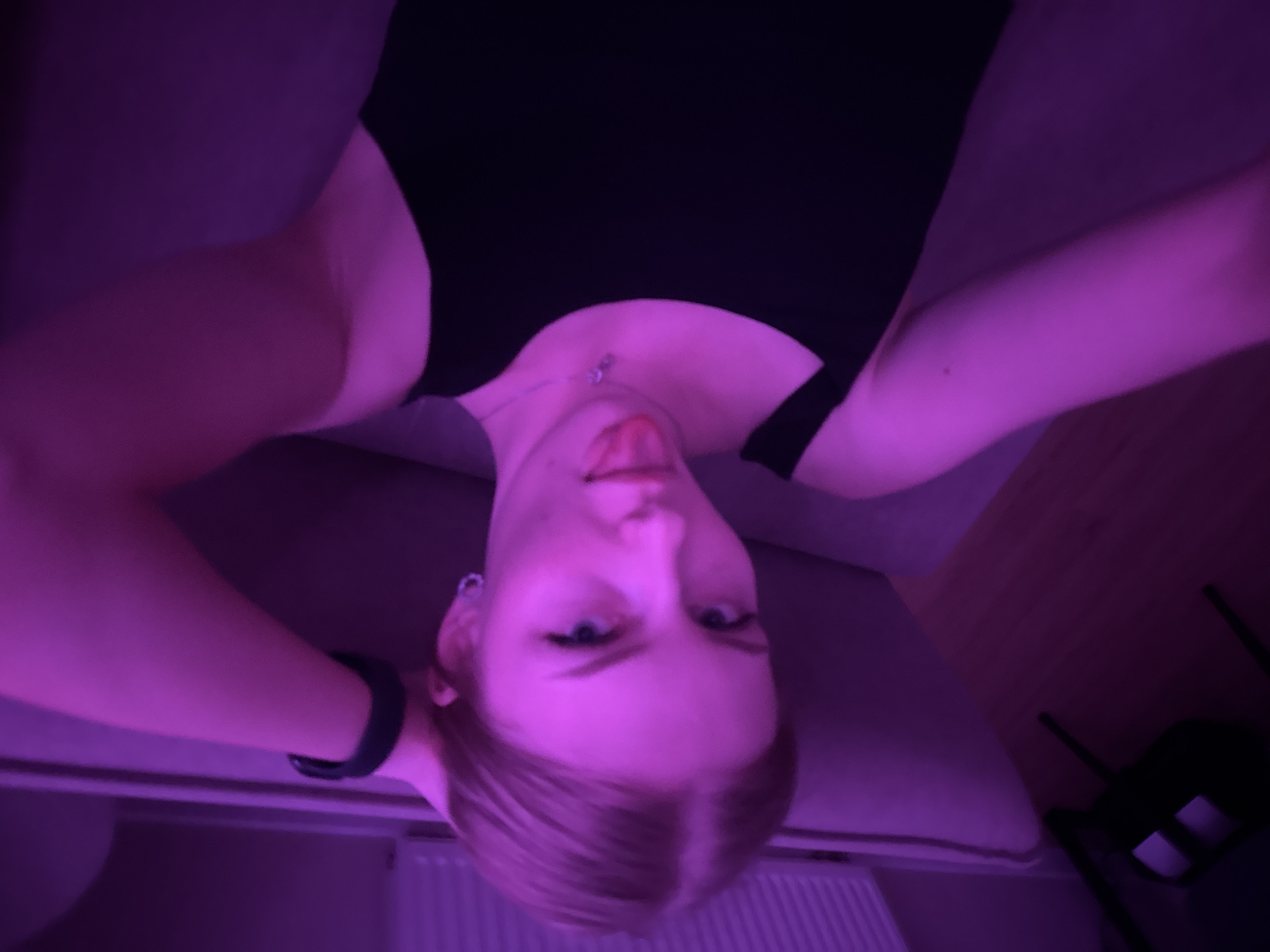 A young woman with blonde hair is lying on a grey sofa, wearing a black top. She is looking at the camera, her head is upside down, and she has her arms behind her head. The scene is illuminated by purple light. In the background, a white radiator is visible, along with a dark chair, possibly made of plastic, and part of a wooden floor. Based on the provided information, the location is potentially Perm, Russia.