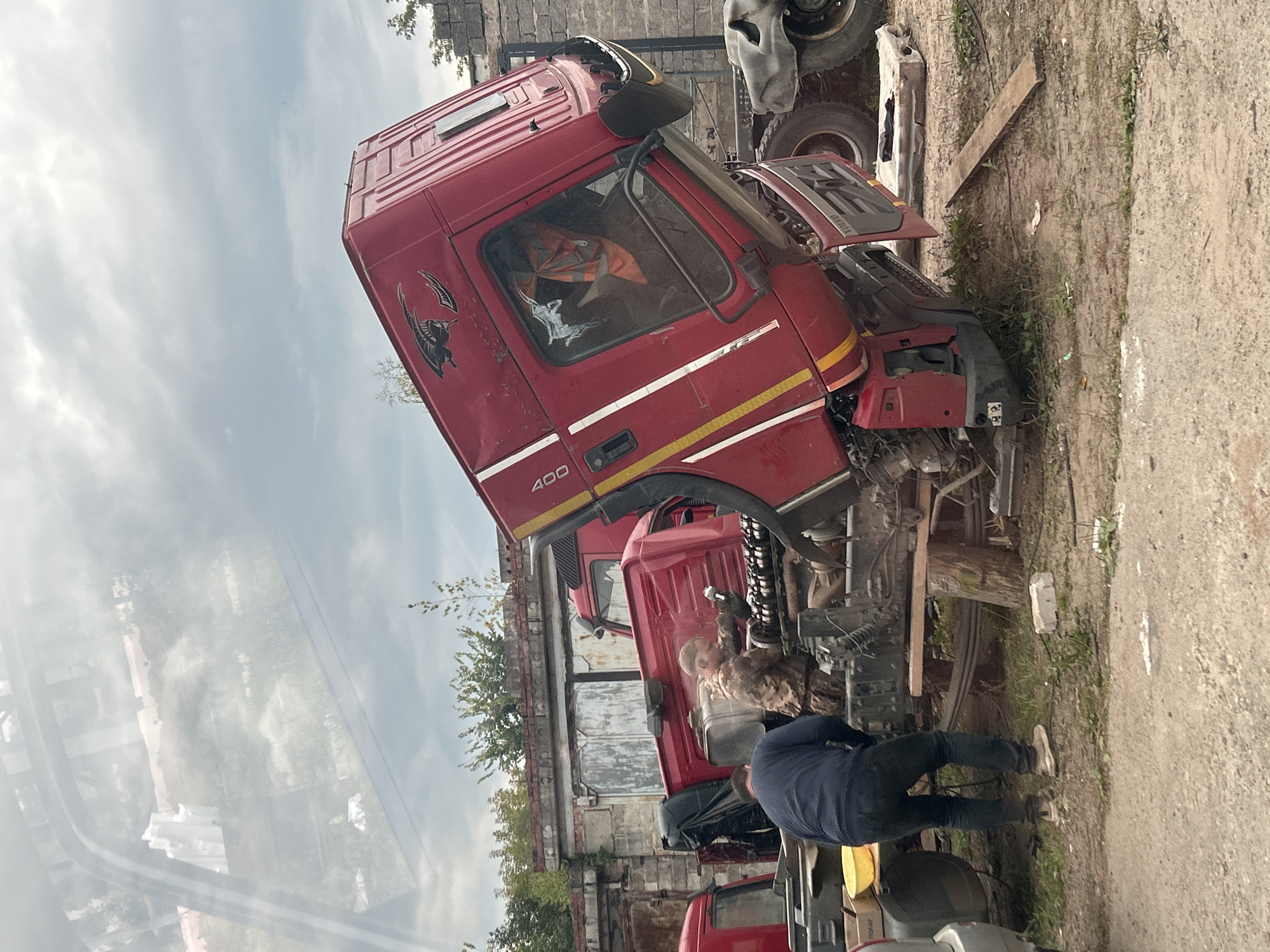 An overturned red Volvo truck is being worked on by two individuals in an outdoor industrial area in Chaikovsky, Russia. The truck cab is tilted at a steep angle, revealing its engine components. One person, wearing a camouflage jacket and pants, is actively engaged in repairing the engine, holding a tool in their right hand. The other person, dressed in a dark blue sweater and jeans, stands near the truck's chassis, observing or assisting. In the background, a brick building with large garage doors and some vegetation is visible. The ground is unpaved and appears to be a dirt and gravel lot. The sky is overcast with clouds. The overall scene suggests a roadside repair or salvage operation.