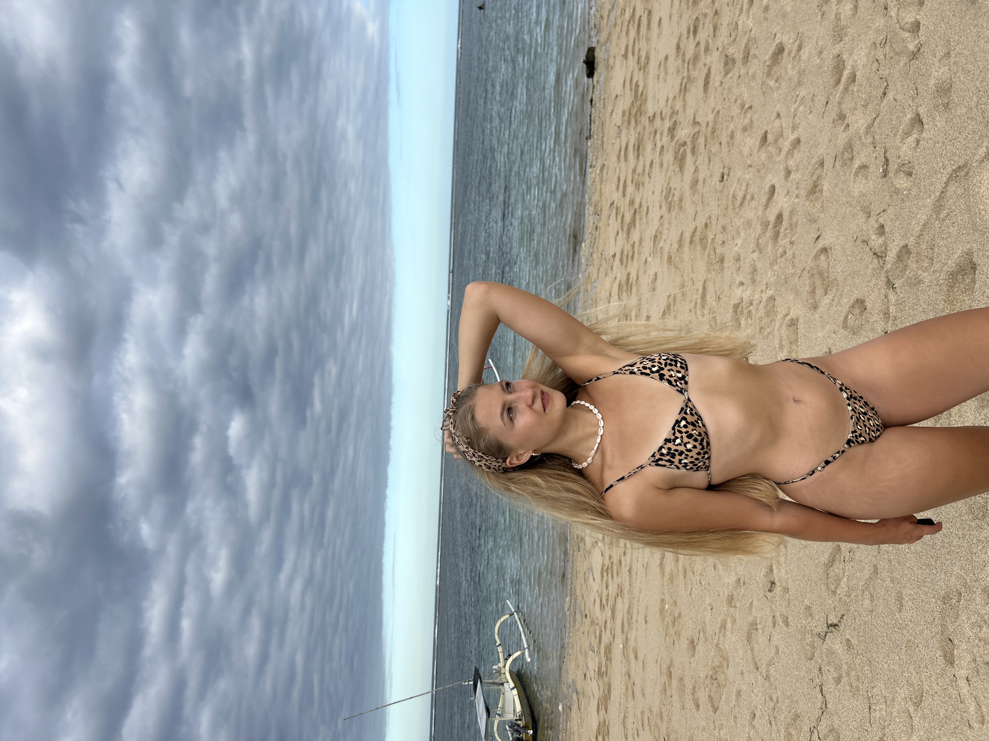 A woman in a leopard-print bikini lies on a sandy beach in Kuta Selatan, Indonesia. Her long, blond hair spreads across the sand. She wears a matching leopard-print headband and a necklace of shells. The sky is cloudy and the water is calm. A small boat sits on the beach nearby.