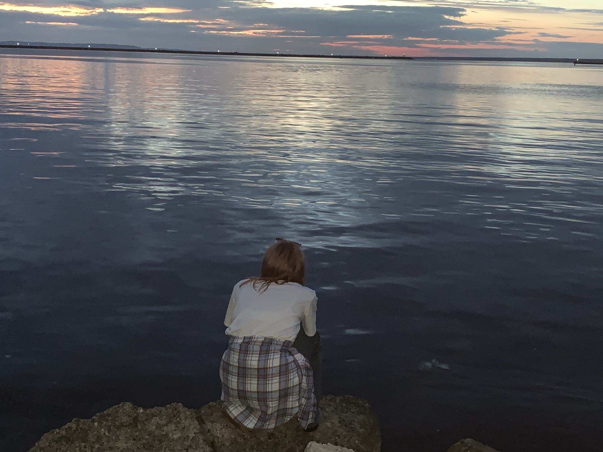A person with reddish-brown hair, seen from behind, sits on a rocky surface facing a large body of water. The person is wearing a white t-shirt and a blue and white plaid shirt tied around their waist. Their legs are bent, and they appear to be looking out at the water. The water is dark and rippled, reflecting the colors of the sky. The sky is a mix of clouds and sunset hues, with streaks of orange and pink. In the distance, a faint landmass is visible on the horizon, with some lights or structures on it. The overall scene suggests a peaceful, contemplative moment by the water at dusk. The location is the Kama River embankment in Chaikovsky, Russia.