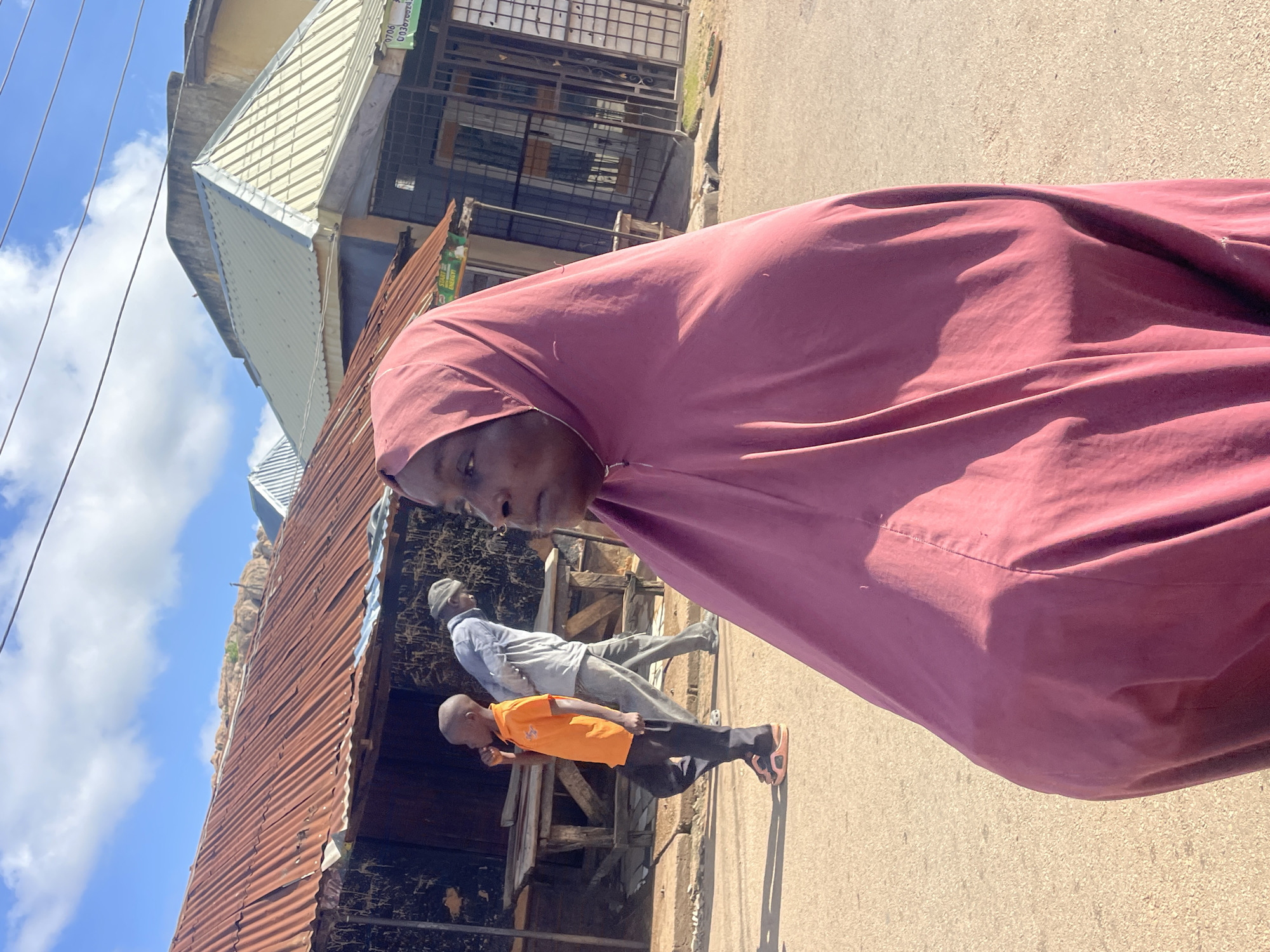 In Jos, Nigeria, a woman wearing a maroon headscarf stands in the foreground, looking to the right. Behind her, a young boy in an orange shirt and dark pants walks away from the camera, while a man in a gray shirt and cap walks in the same direction. The background features a street lined with buildings, including a structure with a rusty corrugated metal roof and another with a lighter-colored metal roof. The scene appears to be a market or commercial area under a bright, partly cloudy sky.