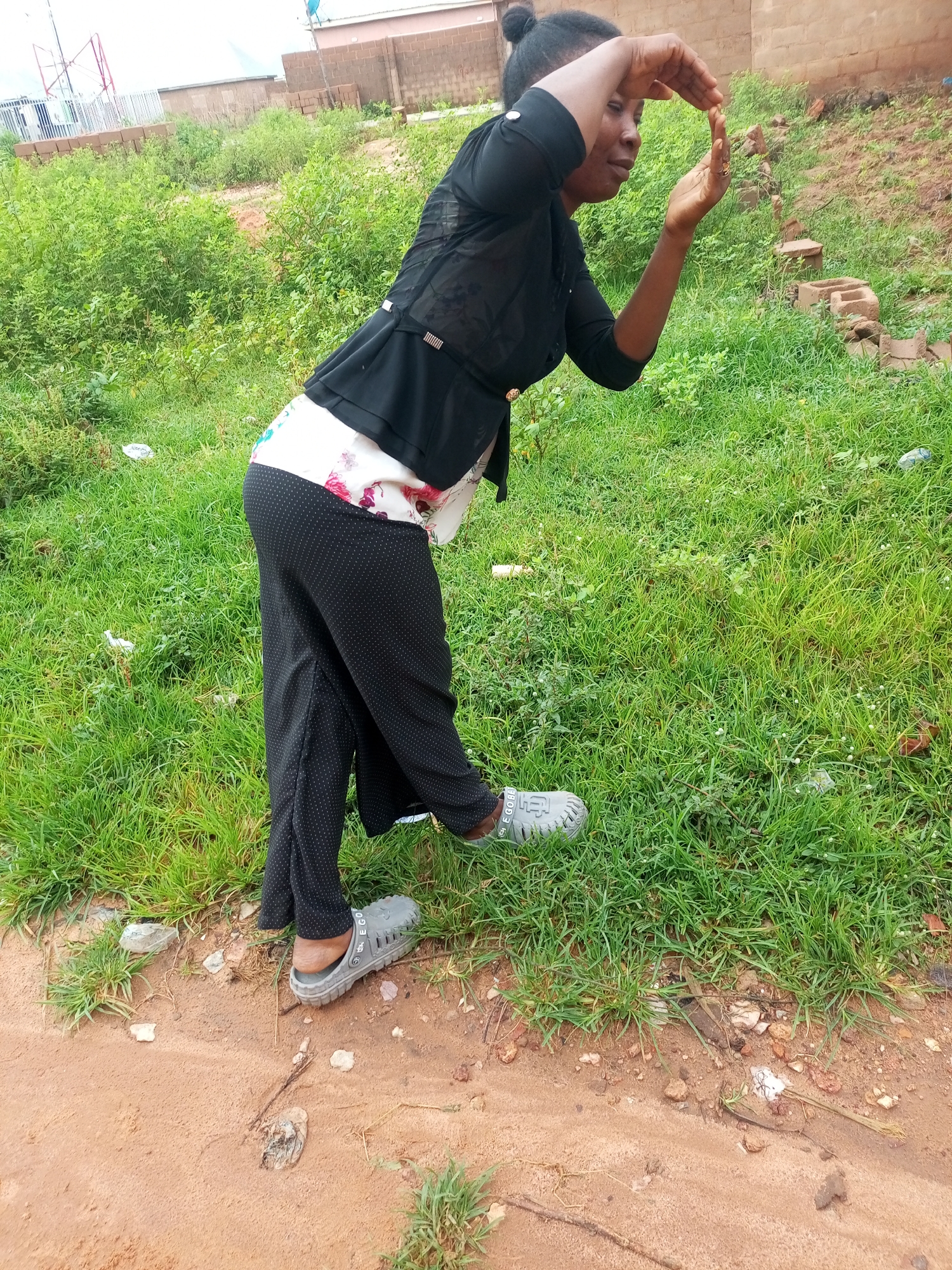 A Black woman is shown from the side, wearing a black shirt with a floral top underneath and black dotted pants. She has her hands cupped in front of her face, as if looking through a frame or holding something. She is standing on a grassy area with a dirt path in the foreground. The background consists of a brick wall and dense green vegetation. The setting appears to be outdoors in a somewhat rural or undeveloped area, possibly on the outskirts of a city like Tempure, Nigeria.