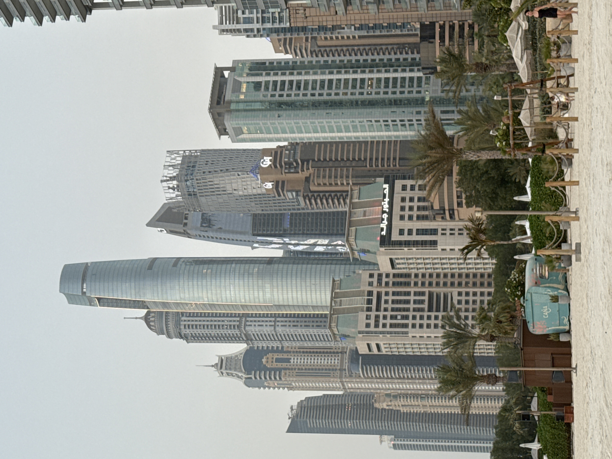 This image depicts a wide shot of the Dubai Marina skyline from a sandy beach. The skyscrapers are tall and modern, with glass facades reflecting the overcast sky. Among them, the tallest building features a distinctive curved design with a lighter section near its peak. To the left, a cluster of buildings includes a prominent structure with a domed top. In the foreground, a beach area is visible with palm trees, lounge chairs, umbrellas, and a small turquoise caravan-like structure with the word "CANA" inscribed on it. A wooden swing set is also present. Several people are scattered across the beach, some walking and others seated, enjoying the coastal environment. The overall atmosphere is serene, with the urban landscape forming a dramatic backdrop to the recreational space.