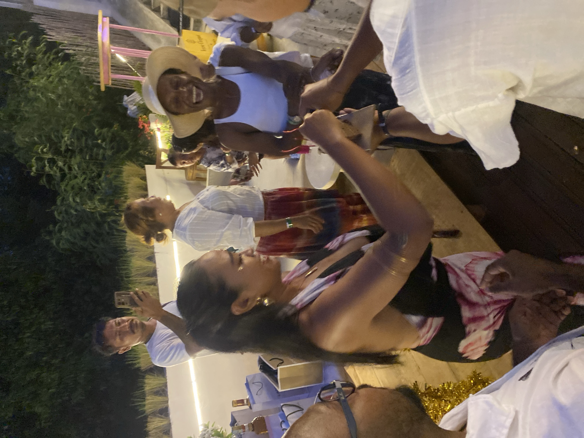 This media file captures a group of people gathered outdoors in a tropical setting, likely in Nusa Penida, Indonesia, as suggested by the location context. The scene appears to be a nighttime event, with warm artificial lighting illuminating the area and creating a festive atmosphere. The ground is a wooden deck, and lush green foliage is visible in the background.
The subjects are primarily adults, dressed in casual, light-colored clothing, consistent with a warm climate. One woman in the foreground, wearing a wide-brimmed hat and a white tank top, is the focal point. She has a wide, joyful smile and appears to be laughing. She is holding an object, possibly a gift or food item, and interacting with another person whose hands are visible. To her left, another woman is seen from the side, also smiling and looking engaged in the activity. Further back, a man is holding up a smartphone, seemingly taking a photo or video of the event.
The overall mood is celebratory and convivial. The event seems to be a social gathering, perhaps a party or a dinner, marked by laughter and shared enjoyment. The presence of gift-like packaging and the interactions between individuals suggest a special occasion.
Notable details include the time of day, which is clearly nighttime, and the warm ambient lighting. The natural elements like the trees and the tropical vegetation confirm the location's likely setting. There is a golden tinsel-like decoration on the ground, adding to the festive decor. A yellow bag with text is visible in the background, but the text is not legible.