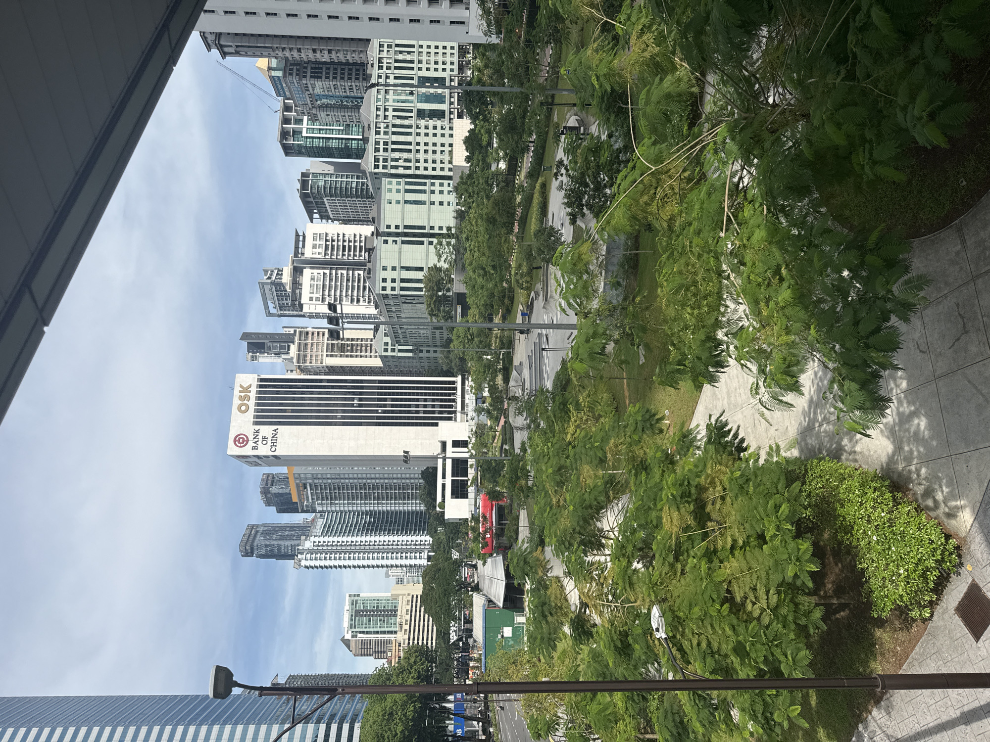 This daytime image, taken from an elevated perspective, captures a dynamic cityscape in Kuala Lumpur, Malaysia, under a clear blue sky with scattered clouds. The scene is dominated by a mix of modern high-rise buildings and lush green urban parkland.
In the mid-ground, a wide road or street is visible with vehicles, including a distinct red double-decker bus, suggesting public transportation or a tour route. Streetlights and infrastructure line the road. To the right, extensive green spaces feature numerous trees and bushes, with paved walkways winding through them, indicating a well-maintained park or landscaped area.
The left side and background are filled with towering skyscrapers of varying designs, reflecting contemporary urban architecture. Some buildings appear to be new or under construction, with a construction crane visible against the sky. A prominent white high-rise features bold black text: "OSK" at the top and "BANK OF CHINA" with its logo underneath. Another smaller blue sign on the street level displays a white "B," possibly marking a bus stop.
The bright lighting and minimal shadows suggest it is mid-morning or early afternoon. The image is devoid of clearly identifiable people, focusing instead on the interplay between the urban environment and its natural green elements.