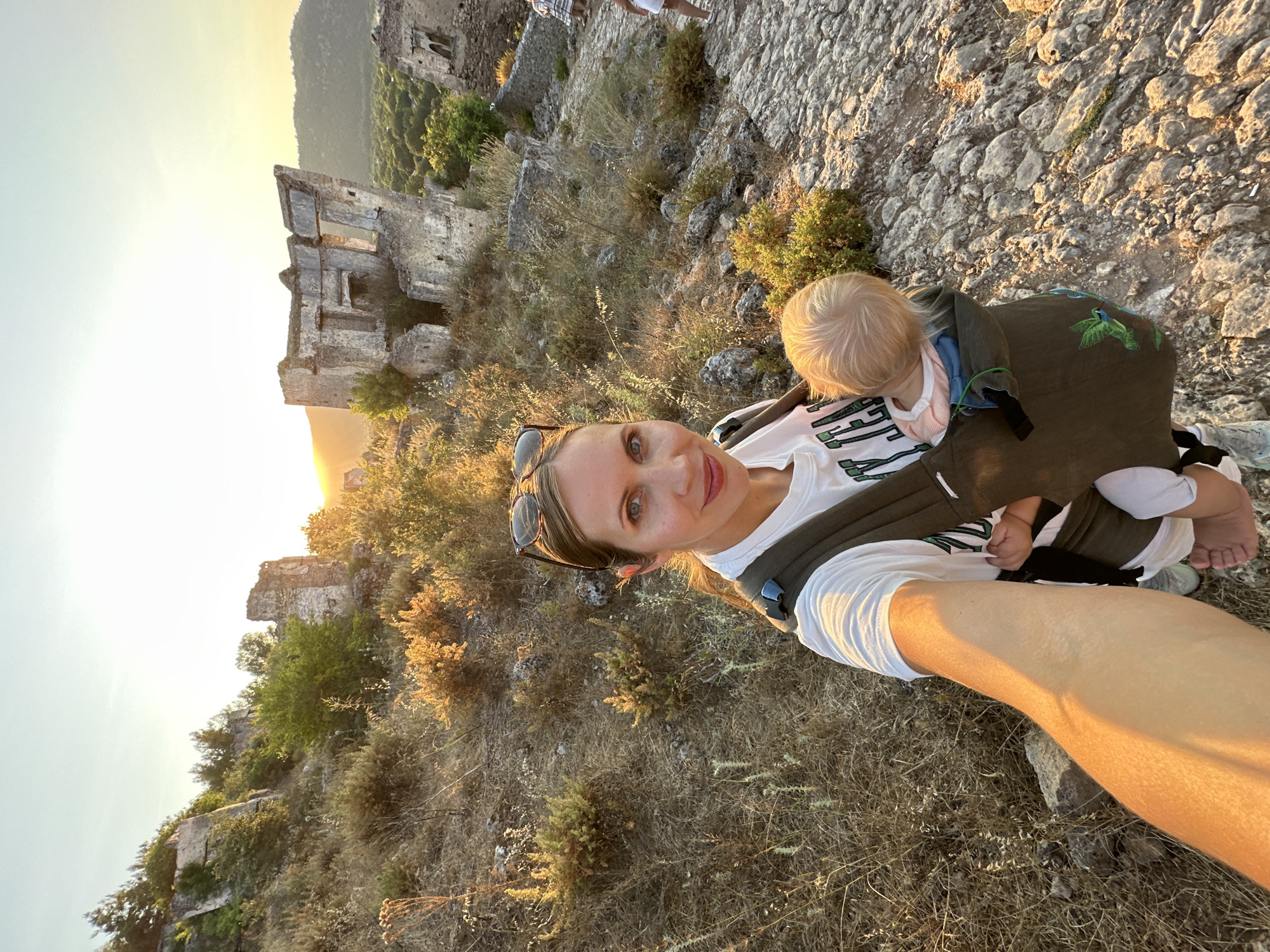 A woman is taking a selfie while carrying a baby in a carrier. The woman has blonde hair and is wearing a white t-shirt and sunglasses. The baby has blonde hair and is wearing a pink shirt. They are standing on a rocky path with ruins in the background. The ruins appear to be ancient stone structures. The environment is dry and arid, with sparse vegetation. The sun is setting, casting a warm glow over the scene. The location is Kayakoy, a ghost village near Fethiye, Turkey.