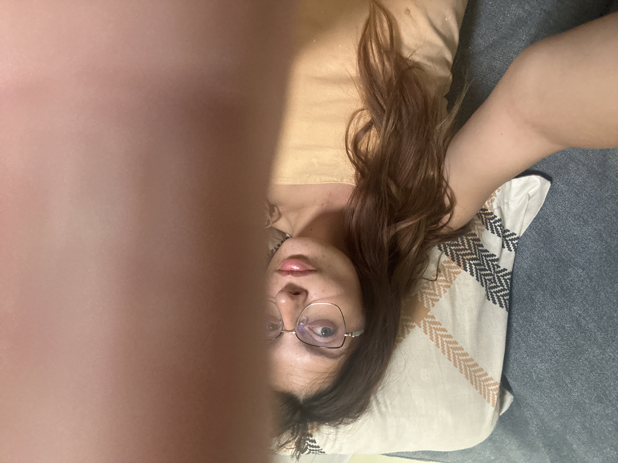 A young woman with long, brown hair and glasses is lying on a sofa, looking at the camera. She is wearing a yellow top. A pillow with a patterned cover is behind her. Part of her arm is in the foreground, obscuring the view. The environment suggests a casual indoor setting, likely a living room. There is no indication of the location being Tryavna, Bulgaria.