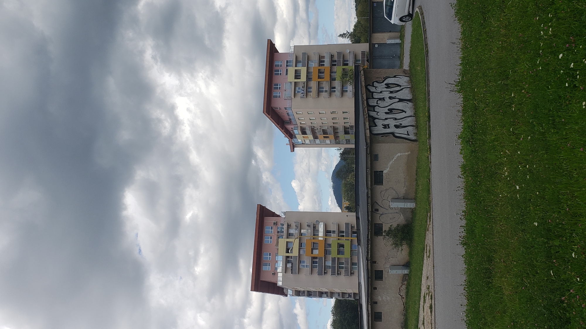 Two identical modern apartment buildings with colorful balconies are visible. A lower, older building with graffiti covers its facade is in the foreground. A white car is parked to the right of the older building. The scene is set against a cloudy sky with patches of blue. The apartment buildings are located in the Sídlisko Sekčov neighborhood of Prešov, Slovakia, specifically near the intersection of Lúčna and Mukačevská streets.