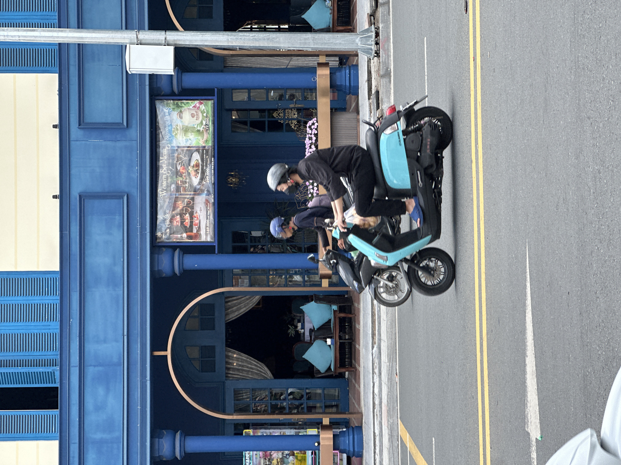 Two individuals on motorbikes are traveling on a street in Da Lat, Vietnam, in front of a vibrantly painted blue building. The building features large, ornate columns, arched doorways, and blue shutters on the upper windows. A banner advertising "Western Set Menus" and "Signature Drinks" is displayed on the building. The street is paved and marked with yellow lines, indicating a traffic lane. The foreground shows the front of a white vehicle, partially visible. The overall scene depicts a typical urban streetscape with elements of local architecture and commerce.