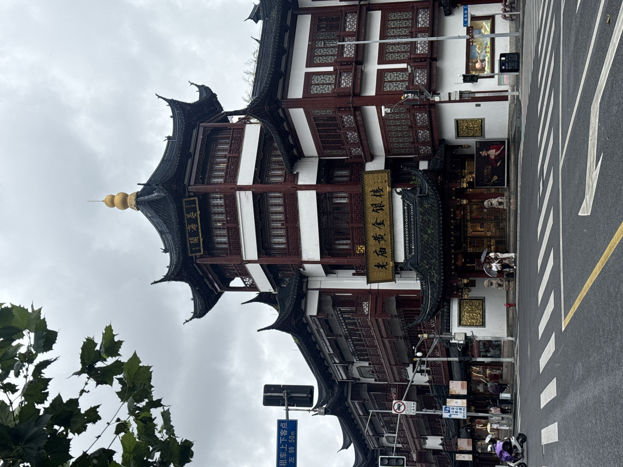 The image captures an overcast day in Shanghai, China, focusing on a traditional Chinese building with multiple tiers and dark, curved roofs. The building is adorned with red trim and white walls, featuring ornate windows. A golden spire crowns the highest roof. Below the spire, a horizontal sign with gold Chinese characters is visible. The building is situated on a street corner, with a pedestrian crossing in the foreground and traffic signals on the left. Several people are present on the sidewalk, some holding umbrellas, indicating light rain. A scooter with a purple canopy is turning at the intersection. Various signs, including a blue directional sign with Chinese text, are mounted on poles. The scene suggests a commercial area with shops.