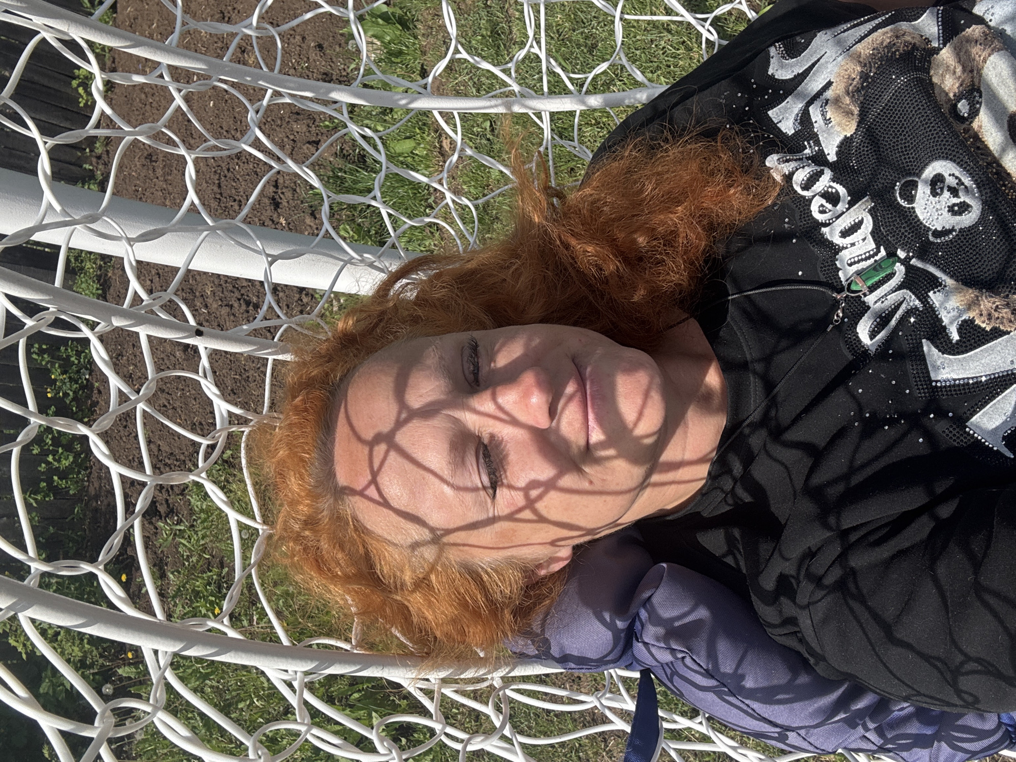 A woman with long, curly reddish-brown hair is depicted resting outdoors, seemingly lying in a white net structure, possibly a hammock or a large net. She is positioned horizontally in the frame, with her eyes closed or partially closed, suggesting she is asleep or deeply relaxed. Strong sunlight filters through the net, casting distinct, intricate shadows across her face and upper body.
She is wearing a black long-sleeved top that features elaborate glittering silver designs. Visible text on the shirt includes part of the word "Super" in large, stylized letters. Cartoonish motifs, such as a panda or bear-like figure and a skull, are also rendered in silver sequins on her top. A green, rectangular pendant hangs around her neck on a thin chain. A blue or purple fabric item, possibly a cushion or backpack, is visible beneath her head.
The background shows patches of green grass and dark brown soil, indicating an outdoor setting, likely a garden or backyard. The bright, direct light confirms it is daytime. The overall scene conveys a sense of peaceful rest under sunny conditions. This image was taken in Chaikovsky, Russia.