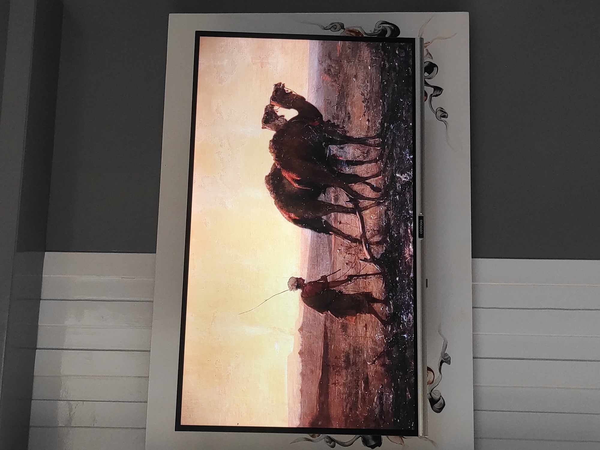 This is a detailed, forensic, matter-of-fact description of the image:
The image captures a framed digital display, likely a television or monitor, mounted on a wall. The display shows a painting of a desert landscape at sunset or sunrise. In the foreground, a person dressed in traditional clothing, appearing to be a man, is leading two camels. The man is holding a long stick or tool, possibly for guiding the animals or for plowing the dry, barren ground. The camels are harnessed together and appear to be pulling something, though the object is not clearly visible. The landscape is depicted with muted earthy tones, with a hazy, warm sky. Decorative elements, appearing as stylized smoky or floral patterns, are present on the white frame surrounding the display. The wall behind the display is divided into vertical panels, alternating between light and dark gray. The lower portion of the wall beneath the display features a white surface with more of the decorative patterns.
Based on the provided information, this scene is located in the city of Sumedang, Indonesia. There are no discernible landmarks or elements within the image itself that specifically pinpoint the location to Sumedang. The description pertains solely to the visual content of the image.