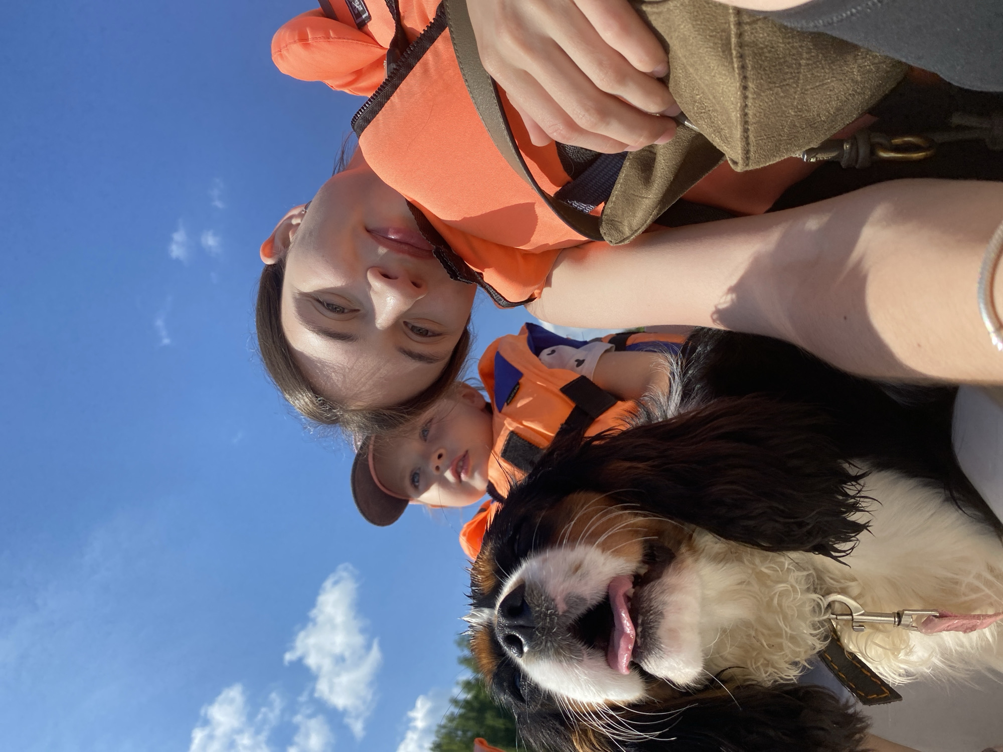 A selfie taken outdoors on a sunny day shows a group in a boat. In the foreground, a dog with black, white, and tan fur is panting with its tongue out. Behind the dog, a young girl wearing an orange life jacket and a brown baseball cap looks upwards. To the right, a young woman with brown hair smiles at the camera, also wearing an orange life jacket. She is holding a brown bag. The sky is blue with scattered white clouds. The image appears to be taken in or near the city of Kamakoye, Russia, as indicated by the context of the image.
