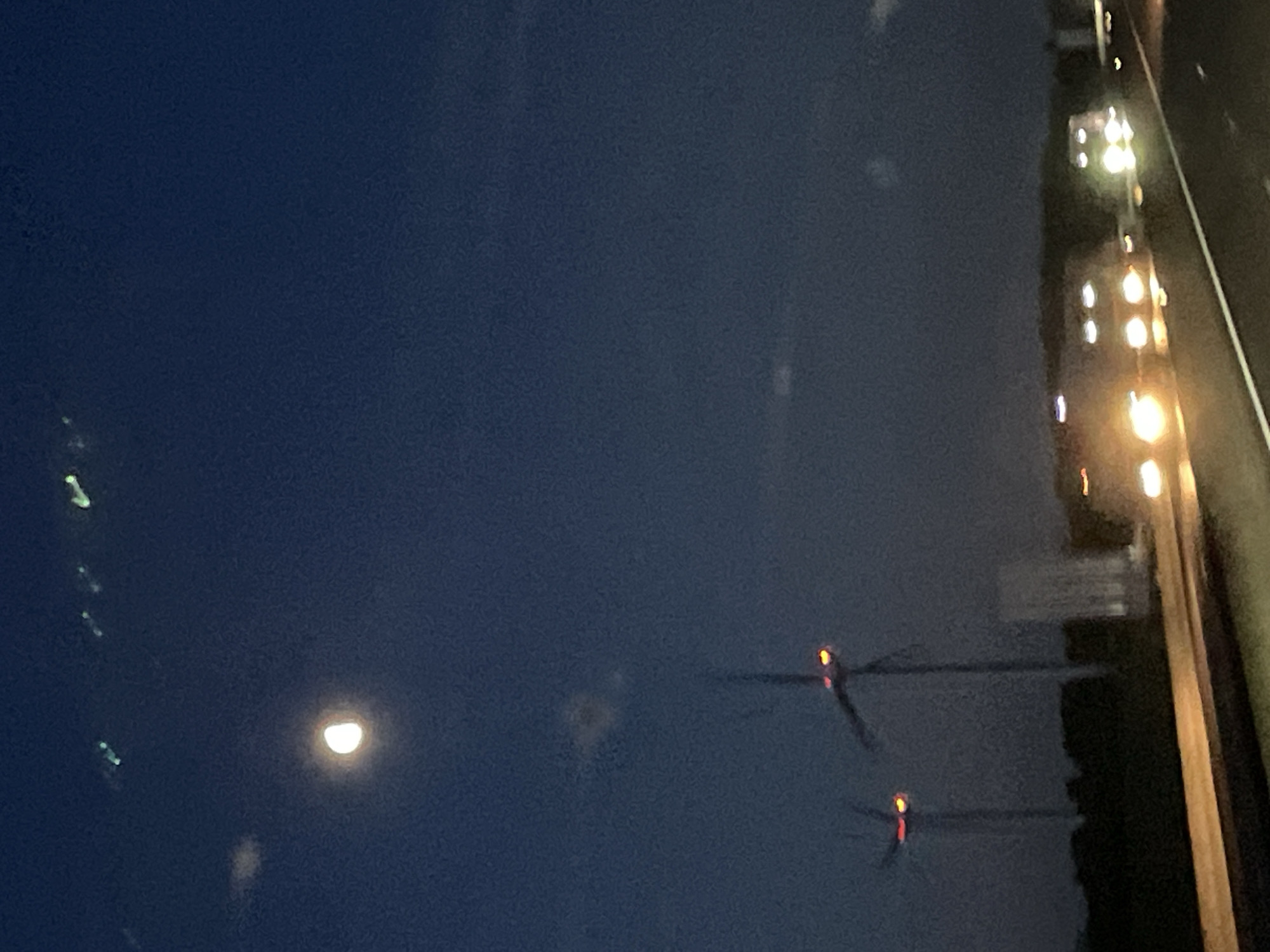 The image shows a dark, nighttime scene, likely taken from a moving vehicle. In the distance to the left, a crescent moon is visible. To the right, a blurry array of lights suggests urban development or a highway with car headlights. There is some evidence of objects that seem like power lines with red markers. The image does not provide sufficient information to place it specifically in the city of Le Val-d'Esnoms, France, nor does it contain any indication of people, specific actions, or interactions. The environment appears to be a roadside setting.
