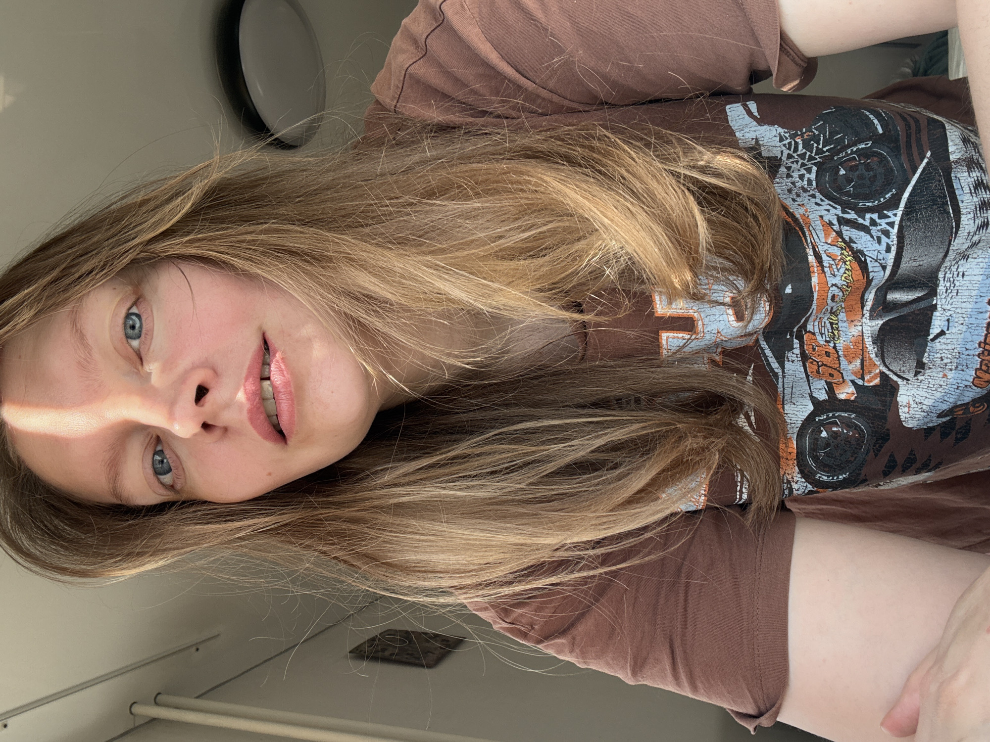 A person with long, blonde hair and blue eyes is pictured. The person is wearing a brown t-shirt with a motorcycle graphic. The background is white with a light fixture visible in the upper left corner. The person is indoors. There is no indication of Izhevsk, Russia.