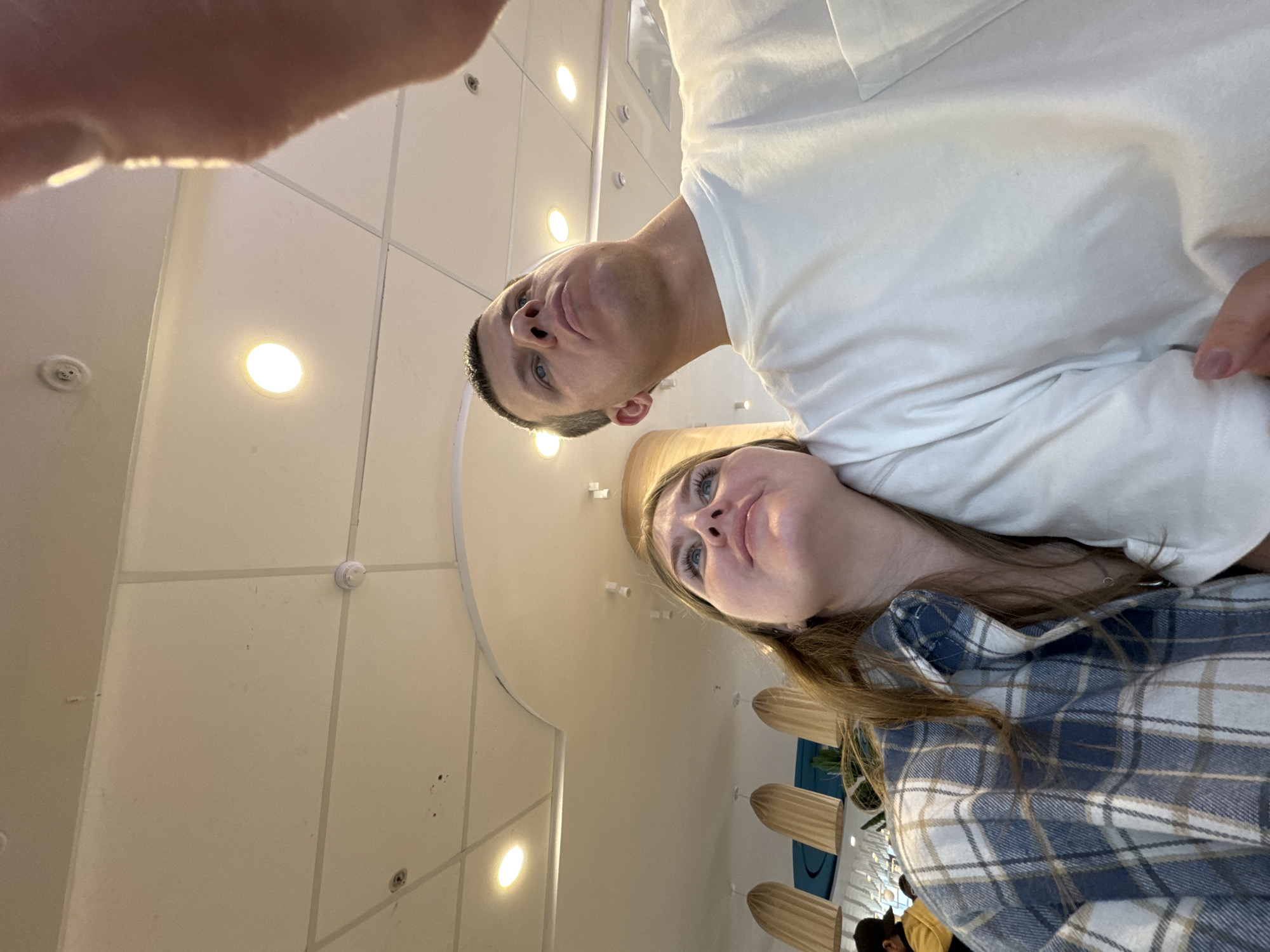 Two individuals are in the frame, a male and a female. The male is positioned above the female, suggesting he is holding the camera. He wears a white t-shirt. The female is wearing a blue plaid shirt.
The background indicates they are indoors, possibly in a public space, evidenced by overhead lighting and ceiling tiles. There are other elements visible in the background such as wooden seats.
There is no indication that the location is in Perm, Russia.