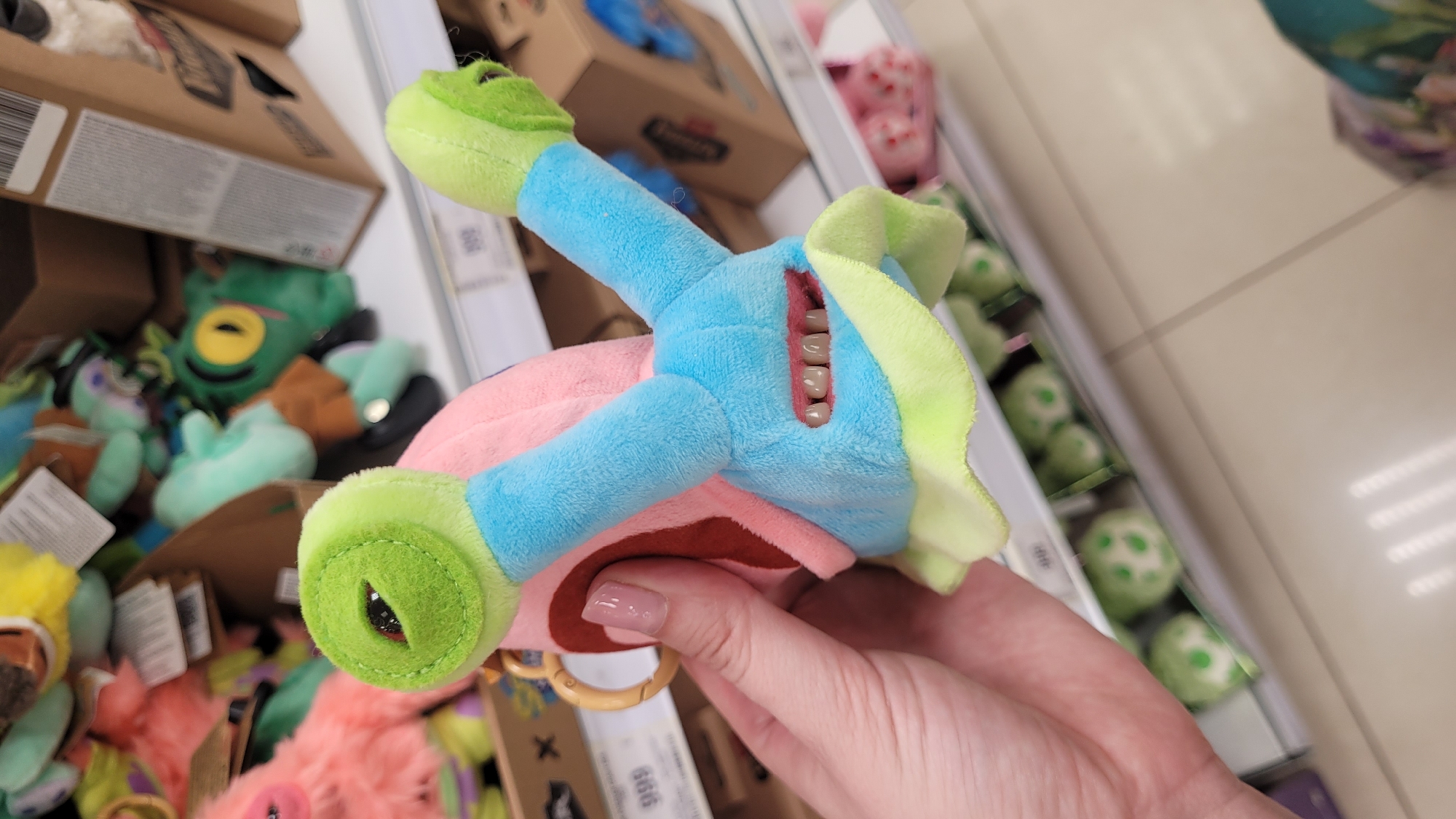 A person holds a plush toy with exposed teeth in a retail environment. The toy is multi-colored with green, blue, pink, and red components. Human teeth are visible in the "mouth" of the toy. The person's hand is partially visible, showing fingernails with light pink polish. The background includes shelves stocked with various other plush toys and retail packaging. The location is a store, in an urban area.