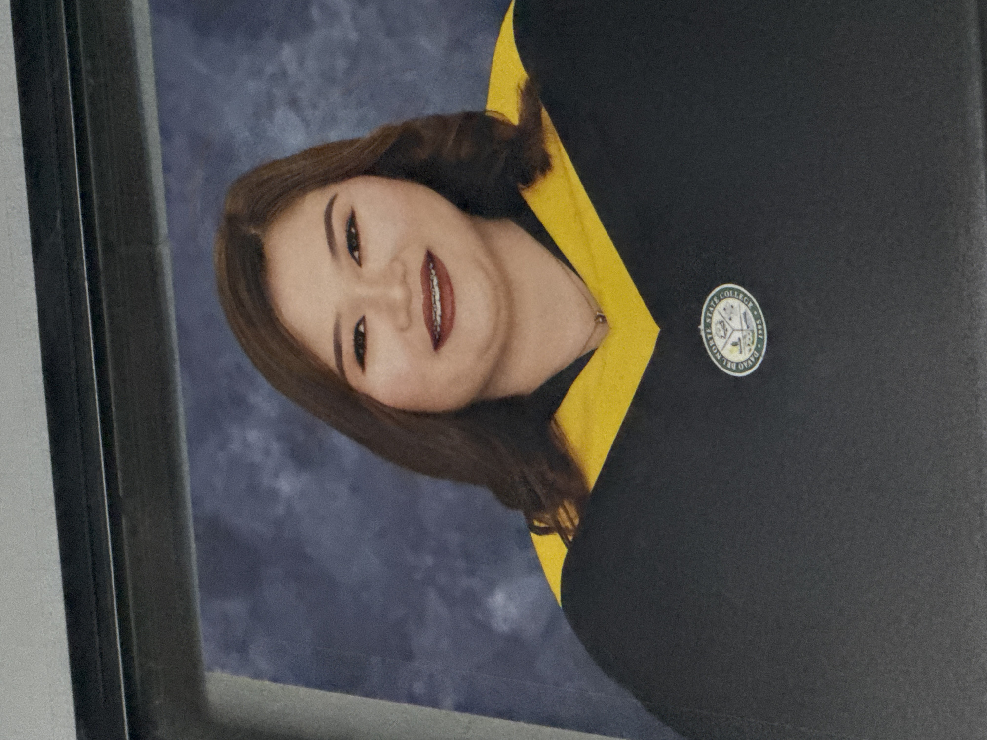 This media file features a formal portrait of a young woman, likely a graduation photograph, in Carmen, Philippines. The image is presented rotated 90 degrees clockwise, depicting a framed picture.
The central subject is a young woman with fair skin, long dark brown hair, and a broad, happy smile revealing dental braces and red lipstick. She is wearing a delicate silver necklace with a small pendant.
She is dressed in a black graduation gown with a prominent yellow trim or shawl around her neck and shoulders. Pinned to the right side of her gown is a circular university seal. Visible text on the seal reads "DAVAO DEL NORTE STATE COLLEGE" and "1995" around its perimeter. Inside the seal, there are symbols including buildings, what appears to be a fish, agricultural stalks, and a sun.
The woman looks directly at the camera, conveying a sense of pride and accomplishment. The background of the portrait is a mottled blue-grey, characteristic of studio photography backdrops. A dark frame is visible along the left edge of the image. The overall scene captures a significant academic milestone.