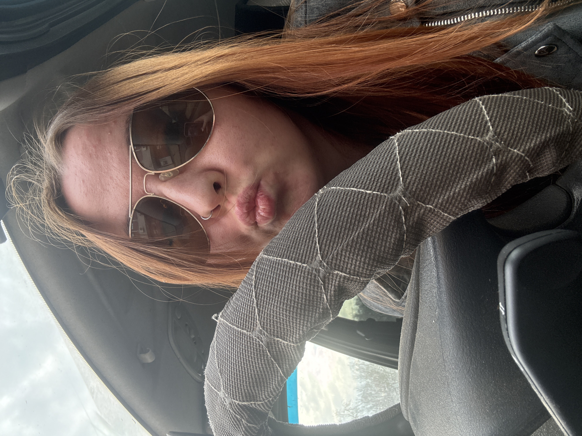 A person with red hair and sunglasses is in a car. The person is positioned behind the steering wheel of the car. The location in the city of Chaikovsky, Russia is not discernable from this photograph.