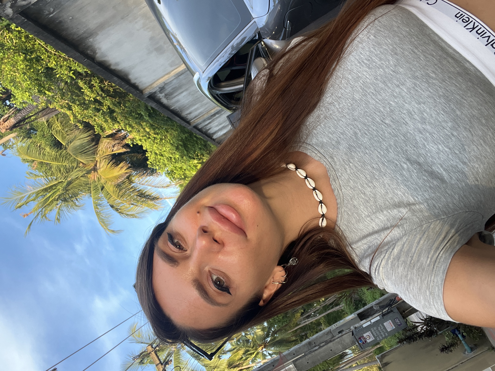 A young woman with long, reddish-brown hair and fair skin is captured in a selfie, looking towards the camera with a slight smile. She has prominent eye makeup, including winged eyeliner, and is wearing a gray t-shirt with a white band that displays "Calvin Klein" in black lettering. Around her neck, she wears a choker necklace made of small white shells. She has several earrings in her left ear, including a hoop and a dangling charm. In the background, lush green foliage, including palm trees, is visible. A concrete wall runs along the right side of the frame, and a portion of a dark-colored car is visible. A utility pole with some electrical equipment and signage is on the left side of the image. The sky above is a clear blue with a few wispy clouds. The overall scene suggests an outdoor setting, possibly in a tropical or subtropical urban area.