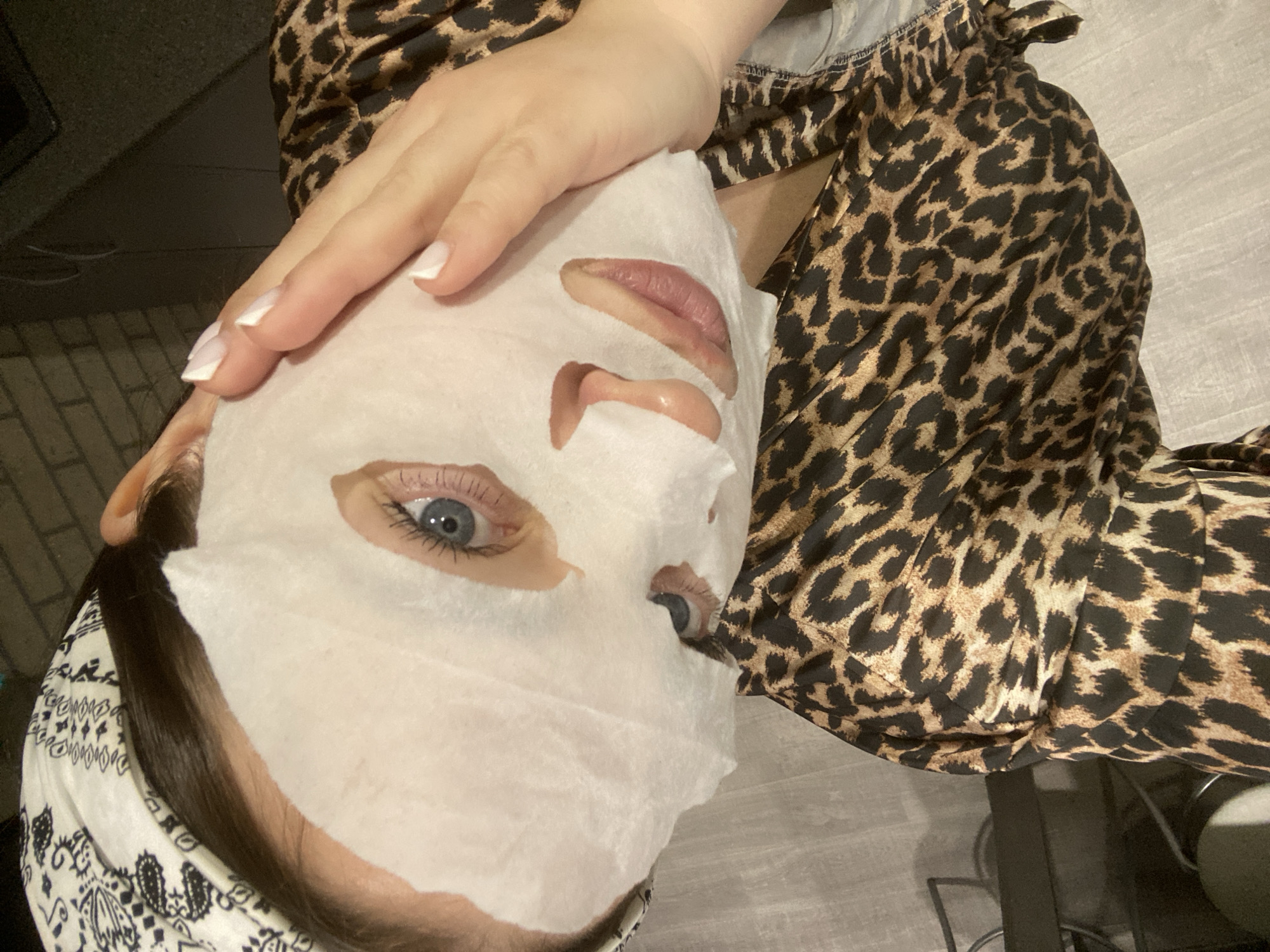 A woman is applying a sheet mask to her face. She is wearing a leopard print robe. Her right hand is on her forehead. She is wearing a patterned hair wrap. The background includes a gray wall and hints of furniture. There is no indication of the location being Sochi, Russia.