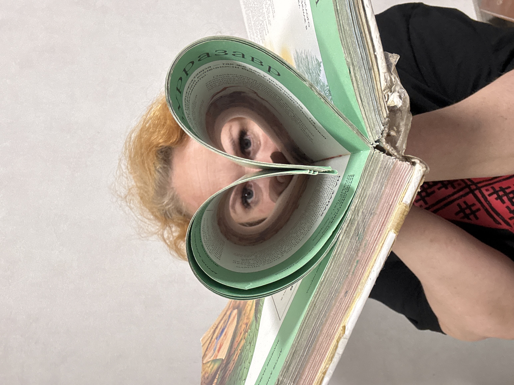 A medium-shot, portrait-oriented photograph depicts a person with light, reddish-blonde hair looking through the pages of a large book. The pages are artfully bent to form a heart shape, through which the person's blue eyes are visible, indicating focus or engagement. The book features green-colored edges on its pages and appears well-used. Cyrillic text is visible on the bent pages, with the fragment "рразавр" clearly legible on the upper left side, suggesting a word like "терразавр" (terrasaur).
The person is wearing a black top, and a section of a red garment with black patterns, possibly geometric, is visible on the right side of the frame. The background is a plain, light-colored wall or surface, indicating an indoor setting. The image is composed at an angle, with the person's head tilted, emphasizing the interaction with the book. The scene conveys a sense of creativity or playful engagement with reading. The location context is Chaikovsky, Russia, though no specific visual cues for this are present in the image itself.