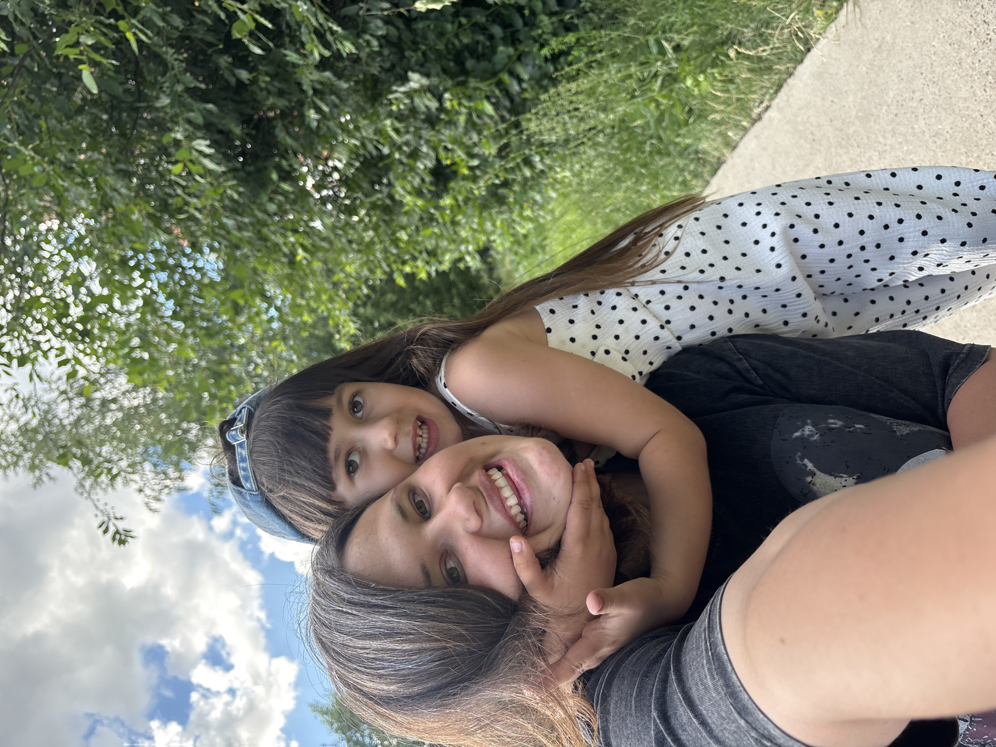 The media file is a close-up, selfie-style photograph featuring two people, likely a mother and daughter, outdoors. The person in the foreground, whose face is partially obscured by the other individual, appears to be an adult with long, dark brown hair and is wearing a grey t-shirt. The person in the background is a child with long, dark brown hair, wearing a white dress with black polka dots and a blue denim headband. Both individuals are smiling and looking at the camera, conveying a sense of happiness and affection.
They are positioned against a backdrop of lush green trees and foliage, with a concrete path visible to the right. The lighting suggests it is daytime, possibly late morning or afternoon, with bright sunlight illuminating the scene. The sky, visible in the upper left portion of the image, is a vibrant blue with fluffy white clouds, indicating fair weather. The overall impression is one of a casual, outdoor outing. No visible text is present in the media file. The location context provided is Chaikovsky, Russia, which aligns with the outdoor, natural setting depicted.