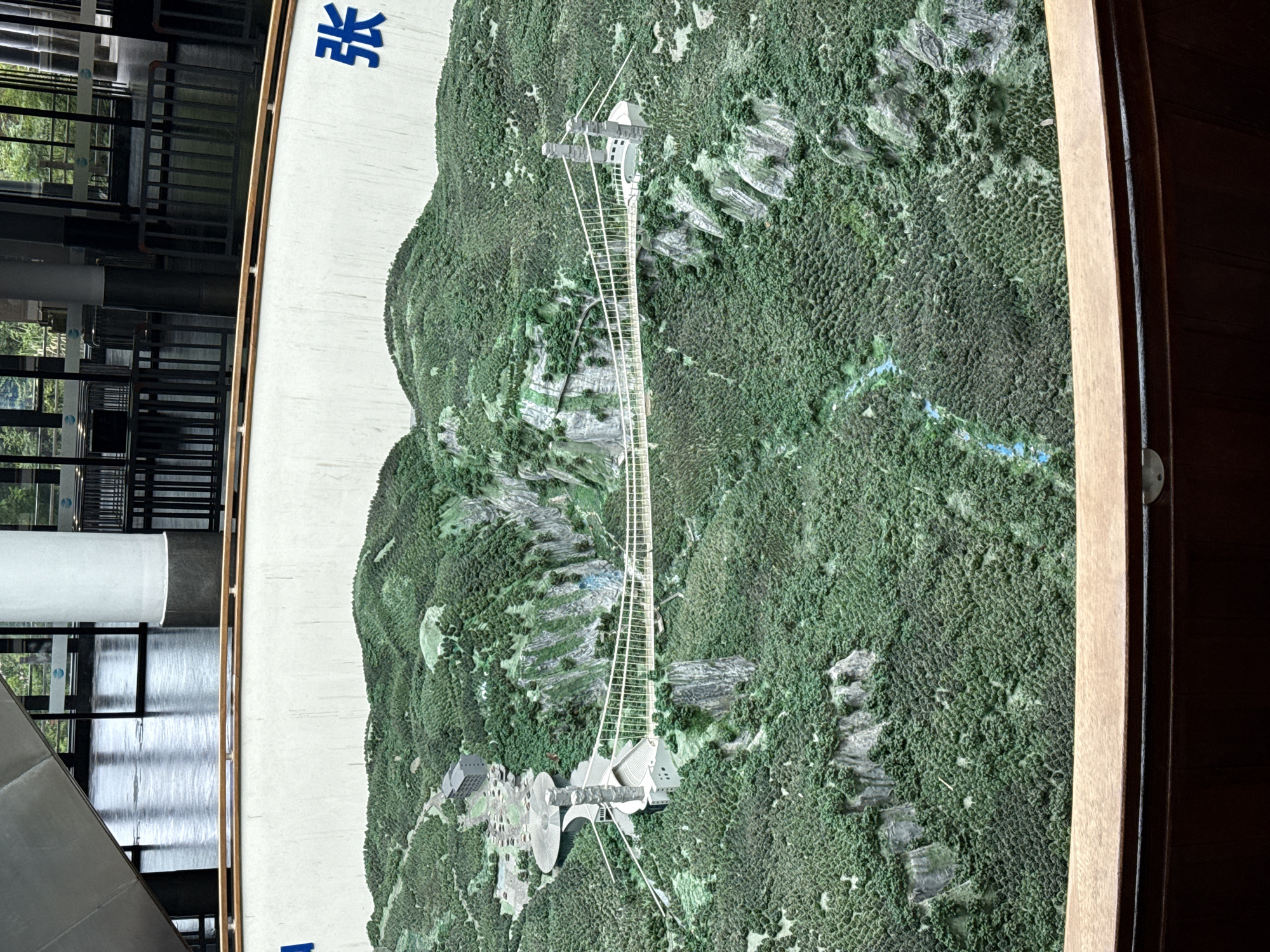 The image displays a physical model of a mountainous landscape, possibly in Zhangjiajie, China, based on the presence of Chinese characters on the model's border. The model shows dense green forest covering the terrain, interspersed with gray rock formations. A long, straight structure, resembling a bridge or walkway, extends through the landscape, connecting two elevated points. One end of the structure leads to a cluster of buildings and circular areas, possibly representing a visitor center or resort complex. A small blue stream is visible amidst the dense greenery. The model is encased in a wooden frame and appears to be on display inside a building, indicated by the presence of a modern structure with glass walls and metal railings visible in the background.