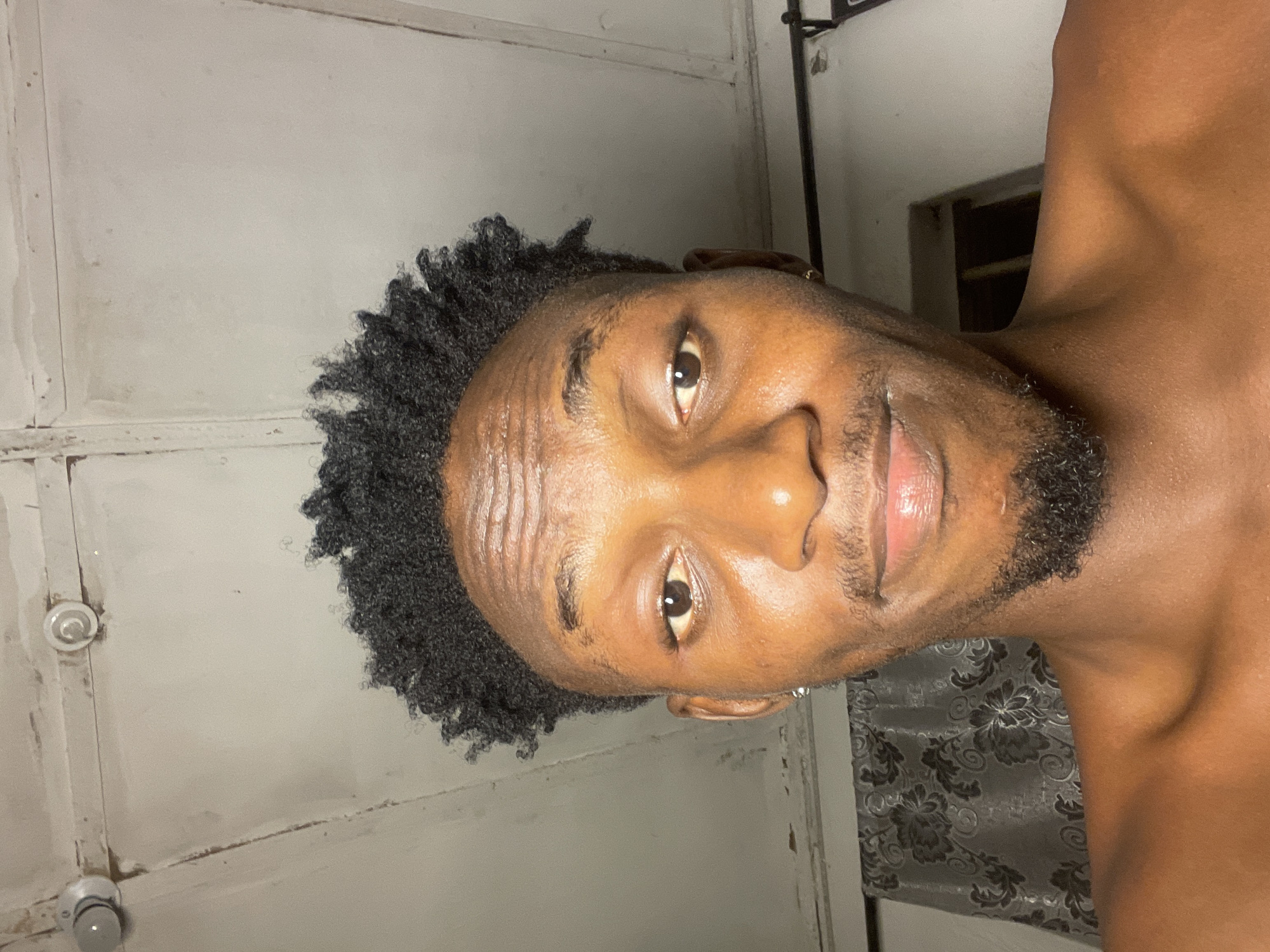 A close-up, slightly tilted portrait of a brown-skinned man with short, dark curly hair and a stubbly beard. He is looking directly at the camera with a slight smile. The background appears to be an interior space with a light-colored wall and a window partially visible on the right side. The location and city of Lokoja, Nigeria cannot be definitively determined based solely on the visual information provided in the image.